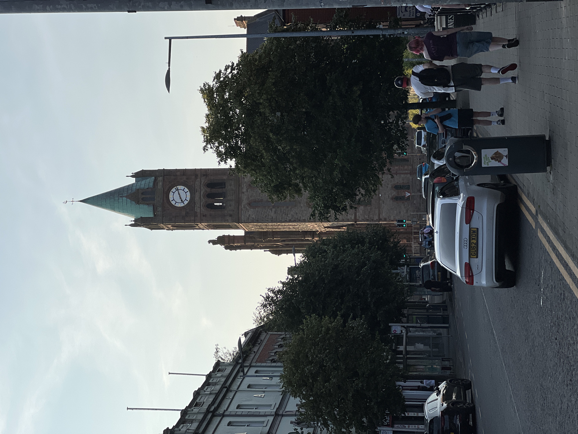 The image shows a street scene in the city of Derry/Londonderry, United Kingdom, with a prominent clock tower, likely St. Columb's Cathedral, dominating the background. The sky is partly cloudy and appears to be late afternoon or early evening.
In the foreground, a paved street runs along the right side of the image, with double yellow lines marking the edge of the carriageway. Several cars are parked on the street, including a white Audi with the license plate GU62 XOW. A pedestrian sidewalk is to the right of the street, with three people walking away from the camera. The person in the middle is wearing a white t-shirt and a backpack, while the person on the right has pink hair and is wearing a dark top and shorts. A young person wearing a blue t-shirt and shorts stands near a trash can.
On the left side of the street, there are buildings with multiple windows and an architectural style that suggests they are older commercial or residential properties. Trees with dense foliage are planted along the sidewalk in front of these buildings. The clock tower, made of brick and with a green spire, rises above the trees and buildings. It features a large clock face with Roman numerals. Streetlights are visible along the sidewalk. The overall impression is a well-established urban environment on a clear day.