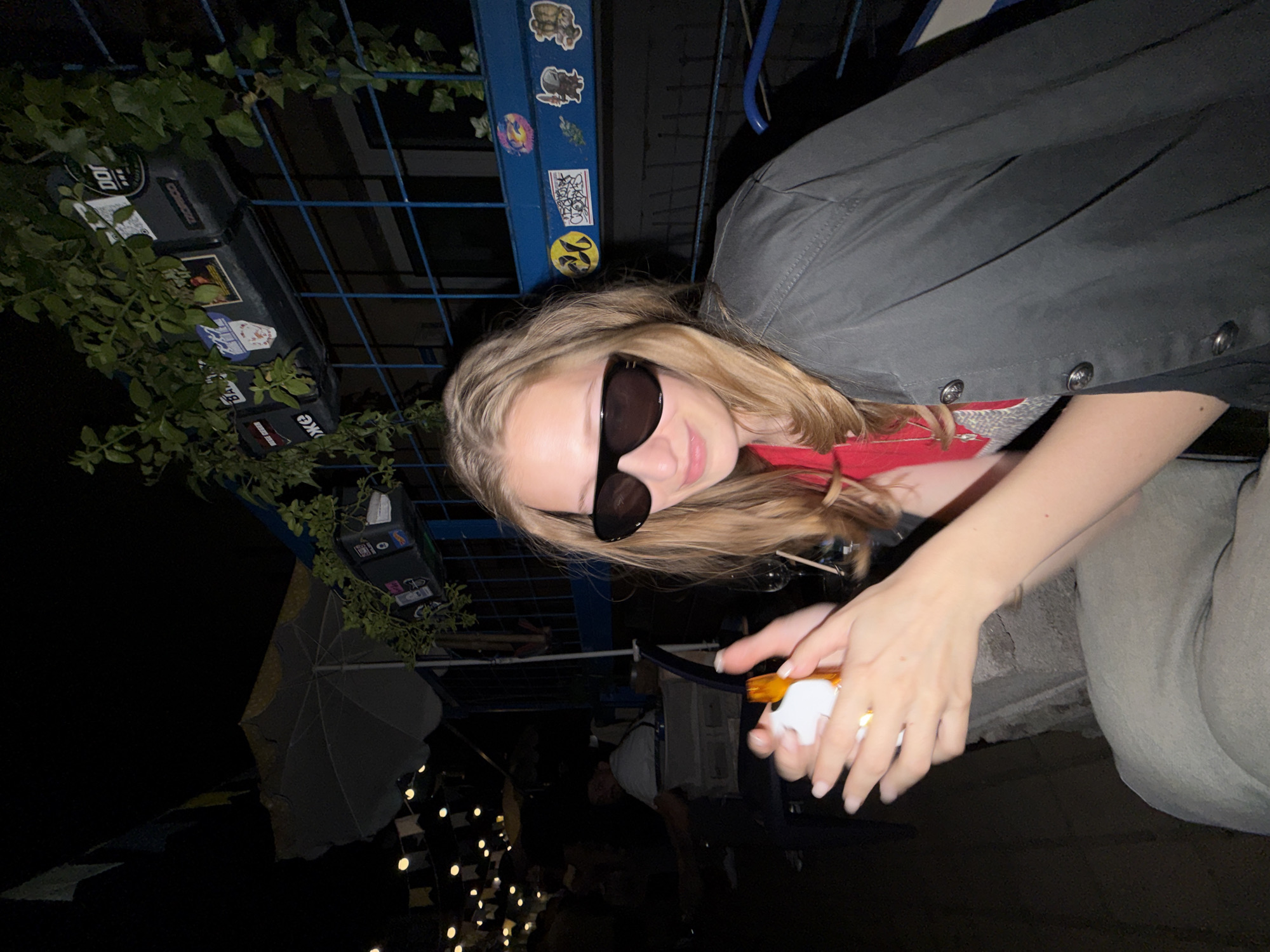 The image depicts a young woman wearing sunglasses and a dark jacket, seated outdoors at night. She is holding a small, white object with an orange tip in her hands. In the background, there are string lights, an umbrella, and a blue railing adorned with various stickers and plants. The setting appears to be an outdoor seating area, possibly a cafe or restaurant. The city of Belgrade, Serbia is not explicitly mentioned or identifiable in the image.