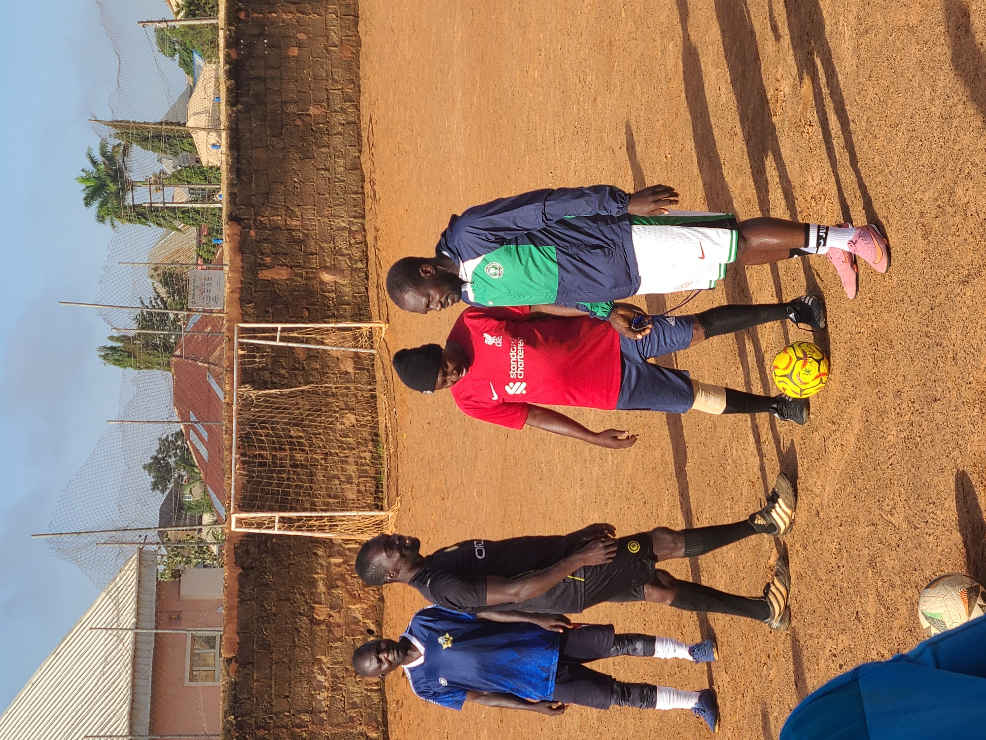 Four men are standing on a dirt soccer field in Barakin Lerawi, Nigeria. The men are dressed in soccer attire. Two men are wearing blue jerseys and shorts, one man is wearing a red shirt and blue shorts, and the fourth man is wearing a blue and green jacket and white shorts. A soccer ball is on the ground between two of the men. There is a soccer goal in the background, and a brick wall behind that. The sky is clear and blue.