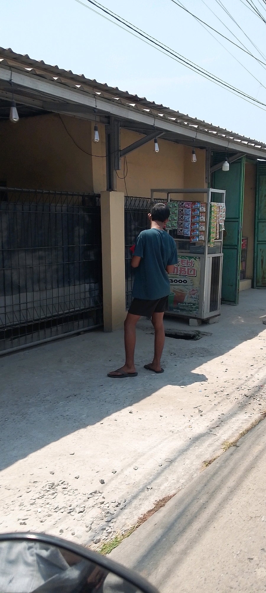 A young male, appearing to be a teenager, stands facing away from the camera in the city of Sukamaju, Indonesia. He is wearing a dark teal t-shirt and dark brown shorts, with brown flip-flops on his feet. The male is standing next to a small, mobile food or drink stand. The stand is made of metal and has shelves displaying various packaged items, possibly snacks or beverages. The stand also has signage, with the words "TEH" and "3000" visible, suggesting tea is sold for 3000 units of currency. Behind the male and the stand is a building with a corrugated metal roof and light-colored walls. A black metal fence is to the left of the male. The ground is paved with concrete and gravel, and the scene is illuminated by bright daylight, casting shadows. Overhead power lines are visible against a clear blue sky. The overall impression is of an outdoor commercial area.