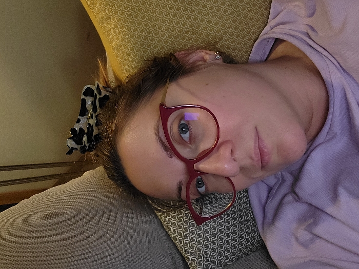 A person with fair skin is lying on a cushioned surface, wearing red glasses. They have blue eyes and pink lips. A black and white patterned hair clip is visible in their hair. The person is wearing a light purple shirt. The background consists of various textured fabrics, with a light-colored patterned cushion behind their head.