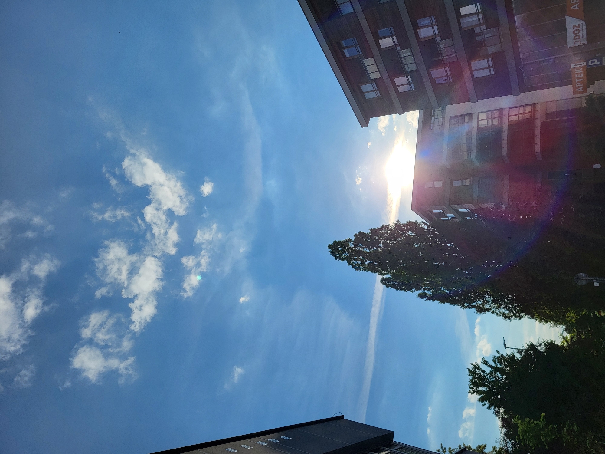 The image shows a view looking upwards. Buildings are visible on the right and at the bottom. On the right, a building with "APTEKA" signage is present. Trees are visible on the right and at the bottom. The sky is mostly blue, with white clouds scattered across it. The sun shines brightly near the building on the right, causing lens flare. The city is Unknown, Unknown.