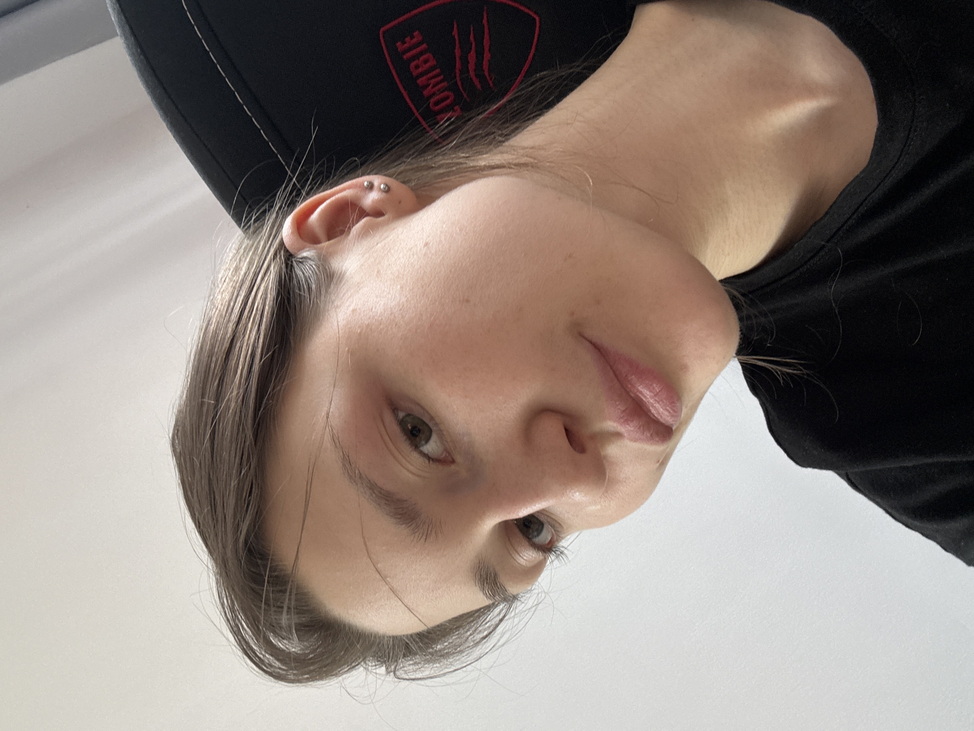 A close-up, tilted headshot of a woman with fair skin, light brown hair, and light brown eyes. She has pierced ears. She is wearing a black shirt and is seated in a black chair with a red logo. The background is a plain white wall. The location is unknown, but no information suggests it is in Saint Petersburg, Russia.