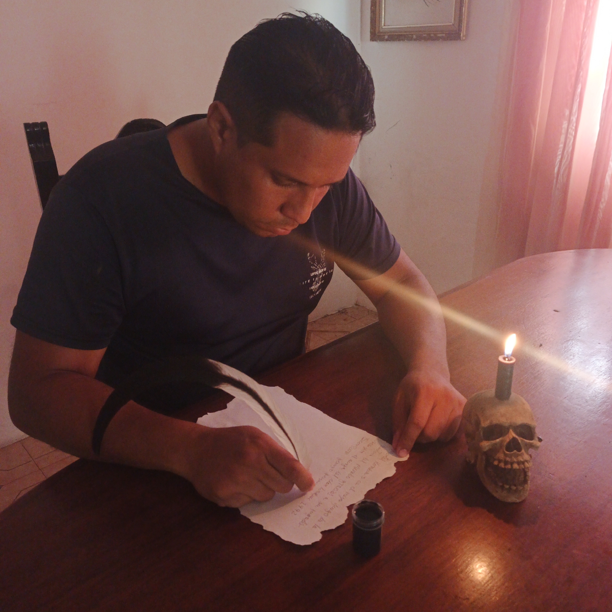 The image depicts an indoor scene located in the city of Tinaquillo, Venezuela. An adult male is seated at a dark wooden table, engaged in writing. He is positioned in the left-center of the frame, with his gaze directed downwards at the writing surface. The male has short, dark hair and is wearing a dark, short-sleeved t-shirt with a light-colored graphic design visible on the left chest area. A solid dark band is present on his left upper arm.
On the dark, polished wooden table directly in front of him, an irregularly shaped piece of off-white paper is laid flat, featuring handwritten text in dark ink that appears to be in Spanish. The male is holding a white feather quill pen in his right hand, actively applying it to the paper, while his left hand rests on the paper to stabilize it. To the right of the paper, a small, dark cylindrical inkwell is present.
Further to the right on the table, a light beige, skull-shaped candle holder, depicting an open jaw with teeth, supports a thin, dark-colored candle. The candle is actively burning, with an upright flame casting a distinct light flare across the table surface and the male's right forearm.
The background consists of a plain, light-colored wall. The upper portion of a dark-colored chair back is visible directly behind the male. In the upper right corner of the background, an ornate, golden-framed picture hangs on the wall. To the far right, a light pink or orange sheer curtain is visible, behind which a bright light source is present, indicating a window or similar opening. A tiled floor is partially visible beneath the table and chair.