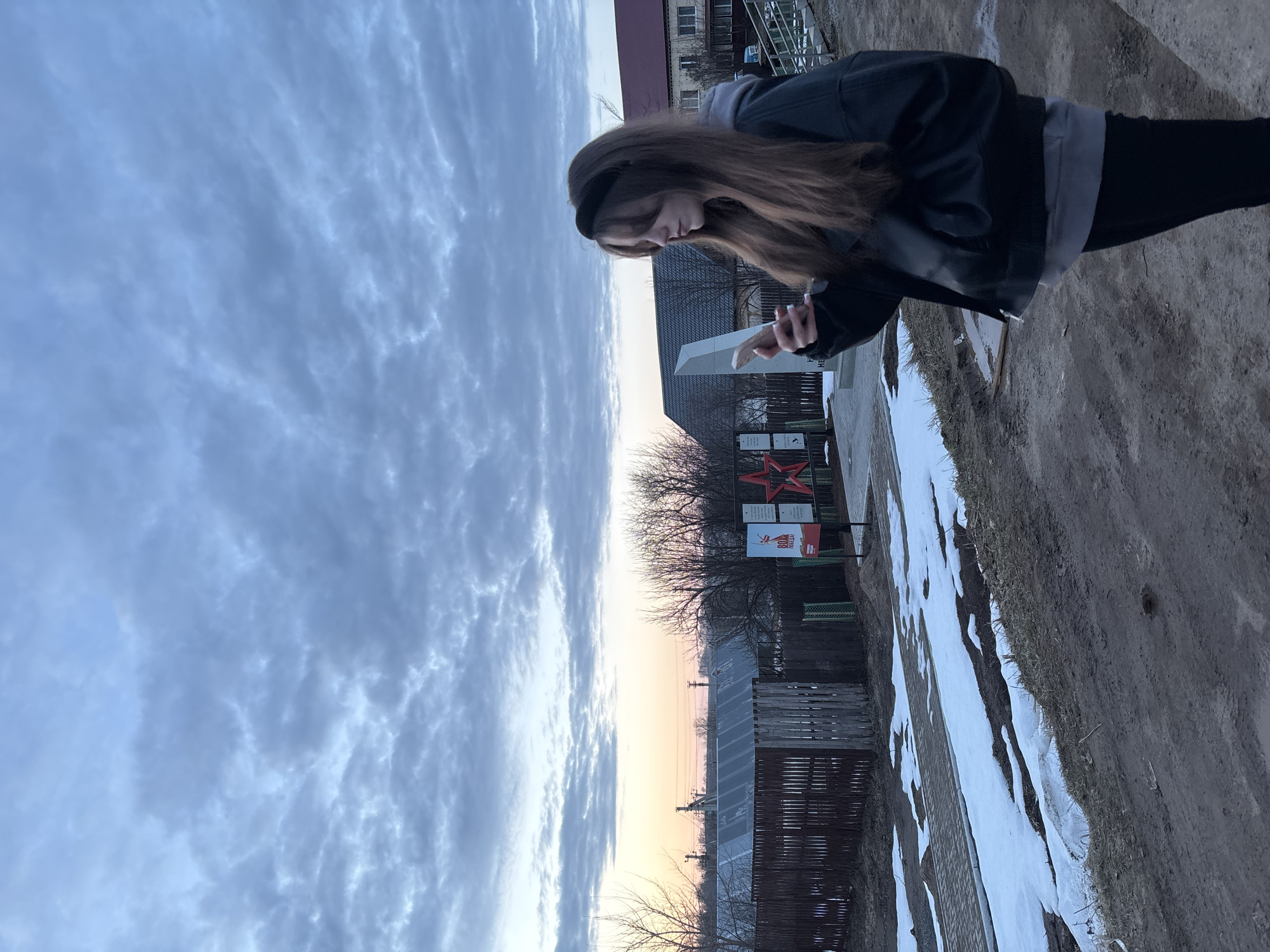A woman is standing outside in the city of Фоки, Russia. The ground is partially covered in snow. A wooden fence is behind her. A monument is behind the fence with a red star on it. The sky is cloudy. The woman is wearing a black jacket, black pants, and a black headband. She is looking at a phone in her hand.