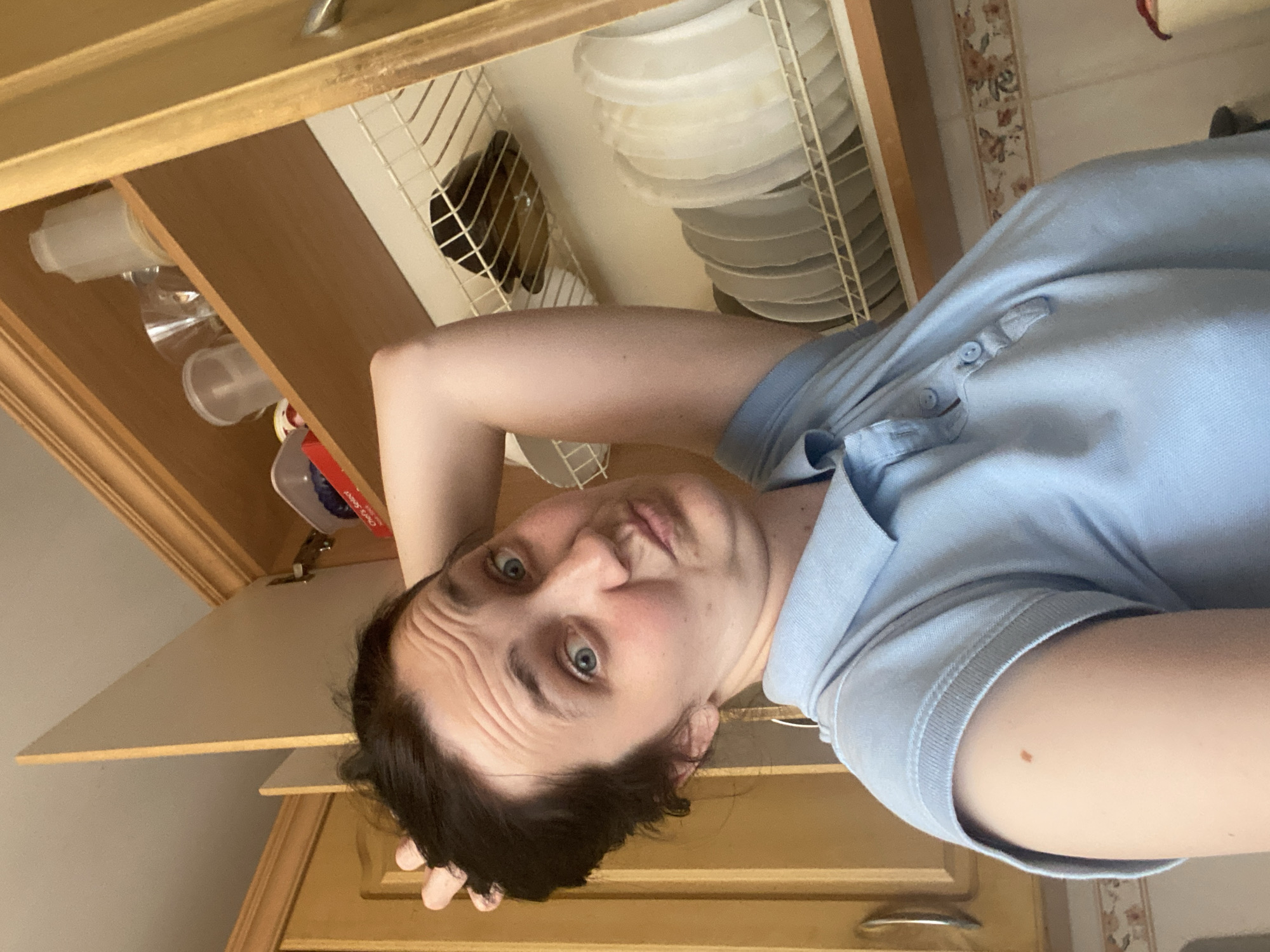 In an indoor kitchen scene, a woman with dark hair is looking at the camera with a playful expression, her right arm raised and hand in her hair. She is wearing a light blue polo shirt. Behind her, an open wooden cabinet reveals shelves containing various kitchen items, including a stack of white plates on a wire rack, and various plastic containers and a glass on an upper shelf. The background shows wooden cabinetry and a floral tiled backsplash. The location is Unknown, Unknown.