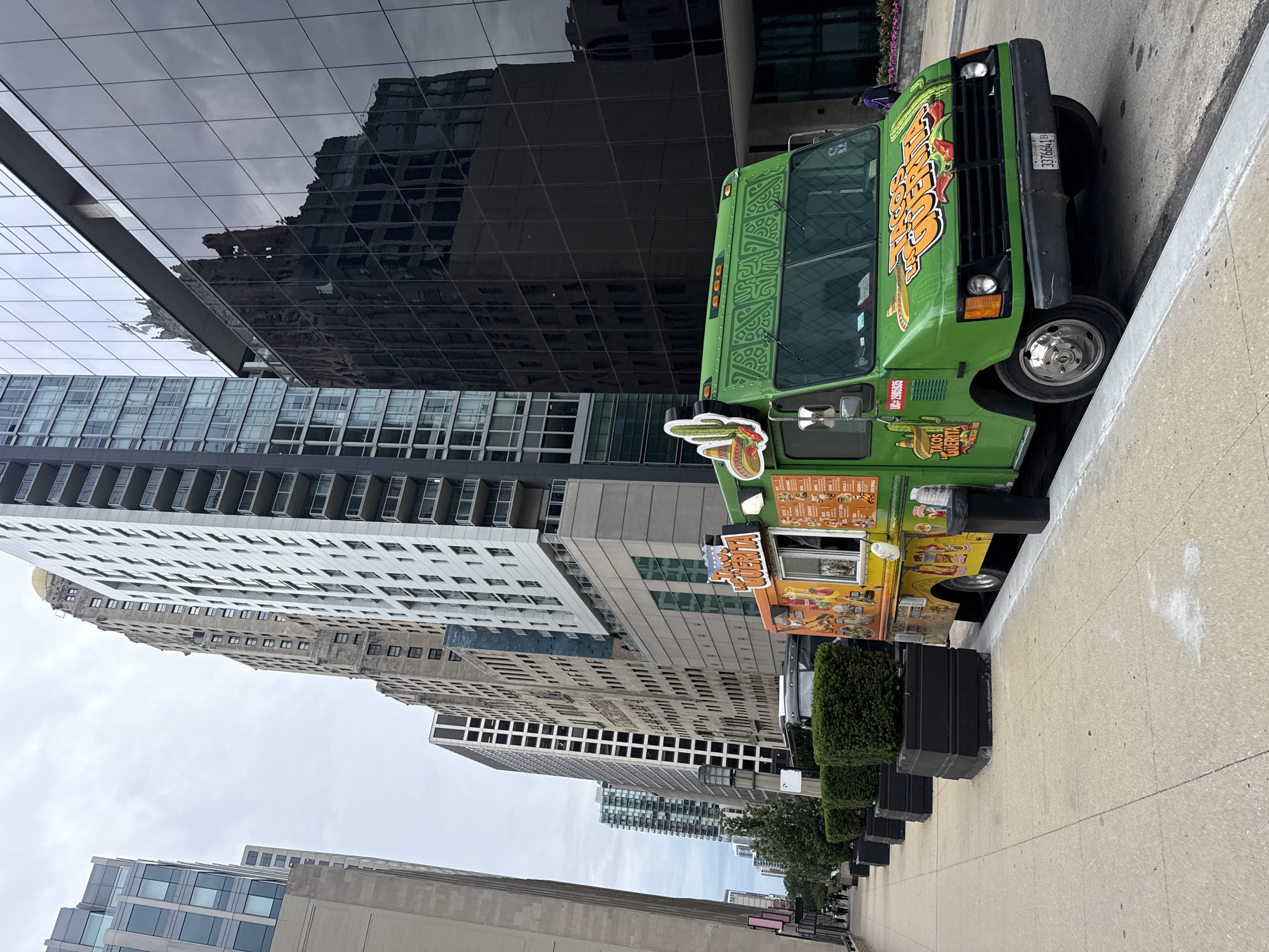 This media file captures a street scene in Chicago, USA, featuring a vibrant green food truck parked on a sidewalk next to a modern building with a reflective glass facade. The food truck, adorned with colorful graphics, appears to be selling tacos and other Mexican cuisine, as suggested by the text "TACOS LA GUERITA" visible on its side. The truck's front displays a cartoonish cactus, and the words "SPICY" and "DELICIOSO" are prominent in its branding. License plate number 3376641B is also visible.
In the background, several tall buildings rise into an overcast sky, creating a typical urban landscape. The reflections in the glass building show other structures, hinting at a densely built environment. The sidewalk is made of concrete, and a row of black planters filled with greenery runs alongside it, suggesting a well-maintained public space.
Although no people are prominently featured, the presence of a food truck implies a commercial activity is likely taking place or has recently concluded. The weather appears to be cloudy, casting a soft, diffused light on the scene. The overall impression is that of a bustling urban environment during daylight hours, perhaps a lunchtime scene where the food truck serves as a popular vendor. The colors of the food truck stand out against the muted tones of the buildings and the sky, making it a focal point of the image.