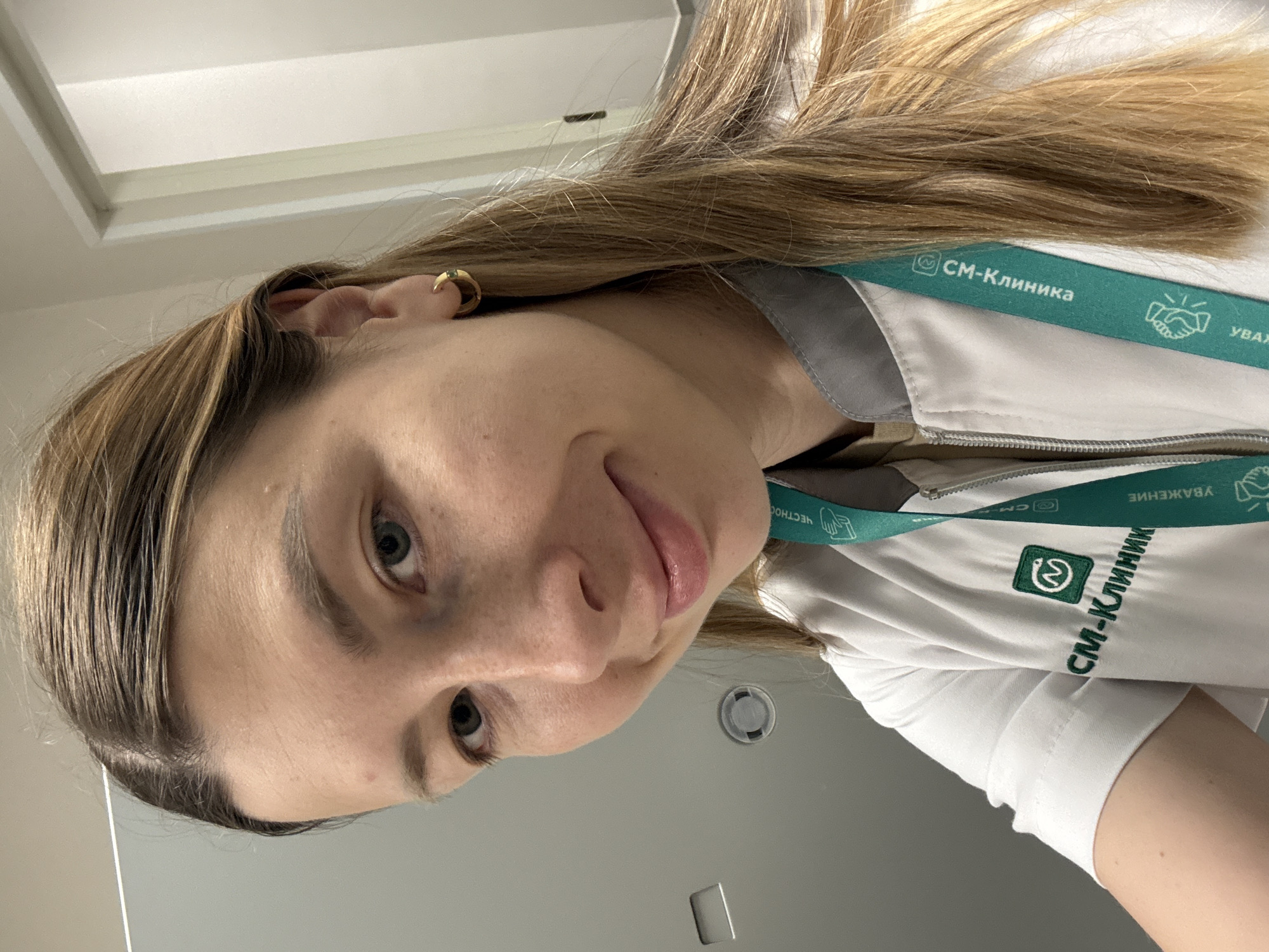 The image shows a close-up portrait of a young woman with long, light brown hair, fair skin, and light-colored eyes. She looks directly at the camera with a subtle, closed-mouth smile or smirk, conveying a casual and friendly expression. She is wearing a white professional top, likely a medical uniform, featuring a zipper and a dark green embroidered logo on the left side of her chest. The logo displays the Cyrillic text "СМ-Клиника" (SM-Klinika).
Around her neck, she wears a green lanyard that also bears the "СМ-Клиника" branding, along with additional Cyrillic words like "УВАЖЕНИЕ" (Respect) and "ЧЕСТНОСТЬ" (Honesty). A small, gold-colored hoop earring is visible in her left ear. The background is simple and light-colored, suggesting an indoor setting with bright overhead lighting, possibly within a medical facility such as a clinic or hospital. The attire and branding strongly suggest she is a medical professional. Given the Cyrillic text, the location is consistent with Moscow, Russia.