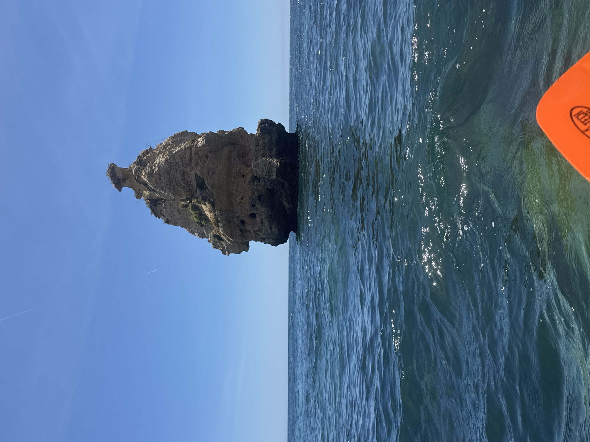 The image shows a coastal scene. A large, rocky island formation is visible on the left side of the frame, contrasting with a clear blue sky above. The right side of the image displays the ocean's surface, rippled with waves and reflecting sunlight. A portion of an orange paddle is visible in the upper-right corner, suggesting the photographer is on a kayak or similar vessel. The location is Unknown, Unknown.
