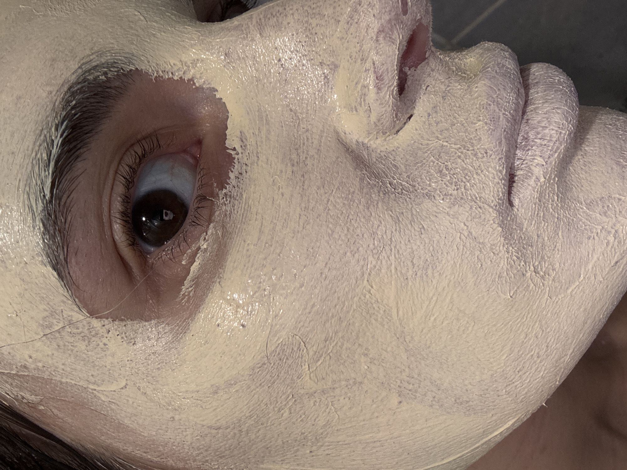 A close-up, low-angle shot focuses on a person's face covered in a pale yellow cosmetic mask. The mask is carefully applied, avoiding the eye area. The person's left eye is clearly visible, with a dark brown iris and visible eyelashes. The mask extends over the forehead, cheeks, nose, and lips. The background appears to be a dark, possibly tiled surface. There's no indication of Chaikovsky, Russia, in the image.