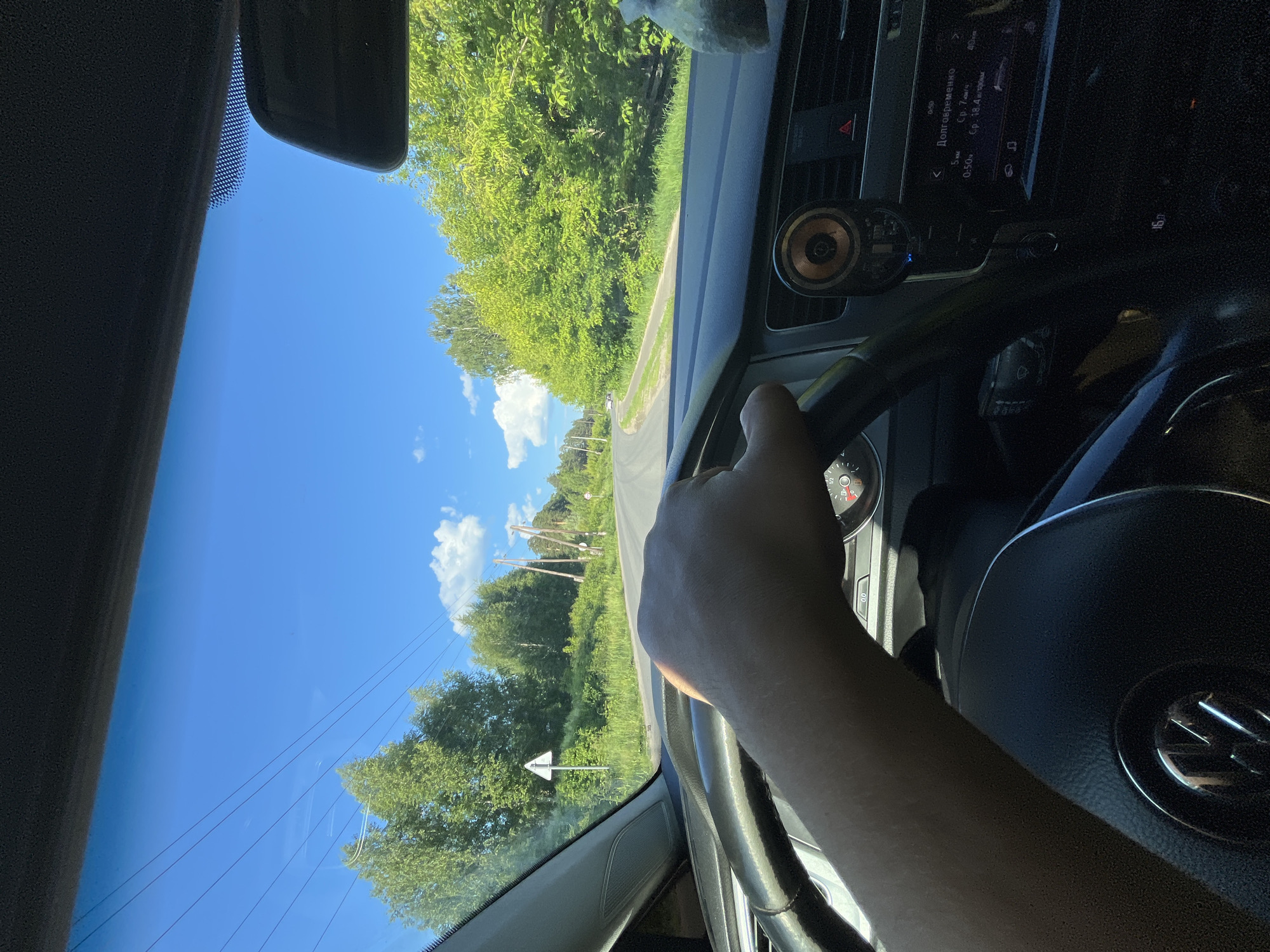 The image is taken from the driver's perspective inside a car, looking forward through the windshield. A person's hand is visible on the steering wheel. The car is on a paved road that curves to the right, surrounded by lush green trees on both sides. The sky is bright blue with some white clouds. Power lines are visible stretching across the sky. There are also some road signs, including a triangular warning sign on the left. In the distance, there are utility poles. The car's dashboard is partially visible, showing a speedometer and infotainment system display. The infotainment screen displays Russian text, indicating the car is likely in Russia. One of the texts reads "Долговременнo" which translates to "Long-term". The presence of Russian text on the car's infotainment system and the road signs suggest the location could be in or near Васильево, Russia.