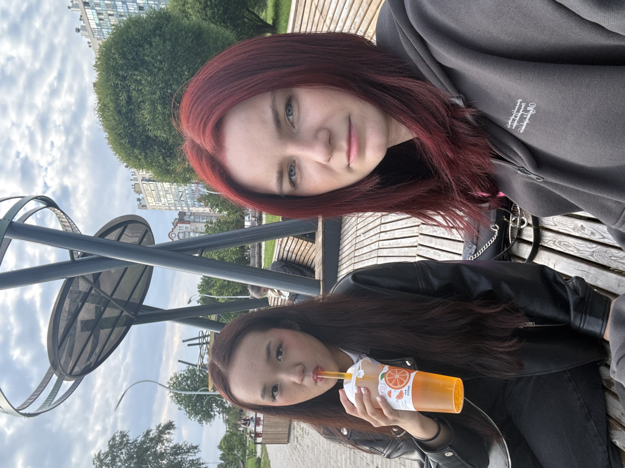 Two young women are sitting on a wooden bench in an outdoor public space, possibly a park, in the city of Unknown, Russia. The woman on the left has long, dark brown hair and is wearing a black leather jacket and black pants. She is holding an orange beverage in a clear plastic cup with a straw and is drinking from it. Her cheeks are slightly flushed pink. The woman on the right has long, dark red hair and is wearing a grey zippered hoodie. She is looking directly at the camera with a neutral expression. The background shows a cloudy sky, trees, modern buildings, and a large, abstract metal sculpture.