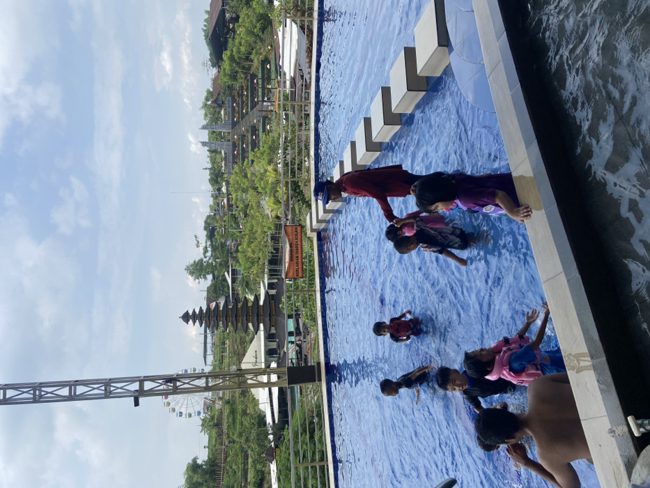 A group of children, some with life vests, are in a blue swimming pool. An adult in a red shirt is assisting them. Behind the pool, there's a lush green landscape featuring a traditional tiered structure, a ferris wheel, and various buildings. The sky is partially cloudy. A metal structure is visible on the left side of the frame, adding a sense of scale to the scene.