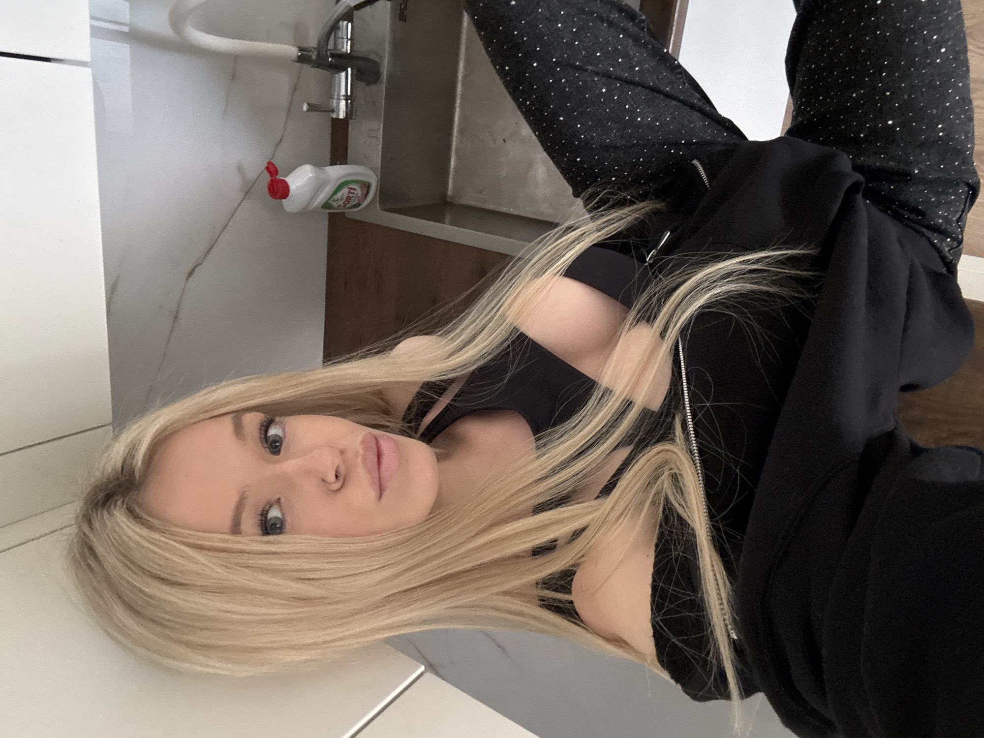 The media file is a selfie of a young woman with blonde hair. She is wearing a black sports bra and black sweatpants with a speckled pattern. A black zip-up hoodie is partially covering her. She is lying down, with her body angled towards the camera. The background appears to be a bathroom or kitchen with white tiled walls and a countertop. A bottle of dish soap is visible on the counter near a faucet. The lighting suggests it could be daytime. The woman's expression is neutral. The location is Saint Petersburg, Russia.