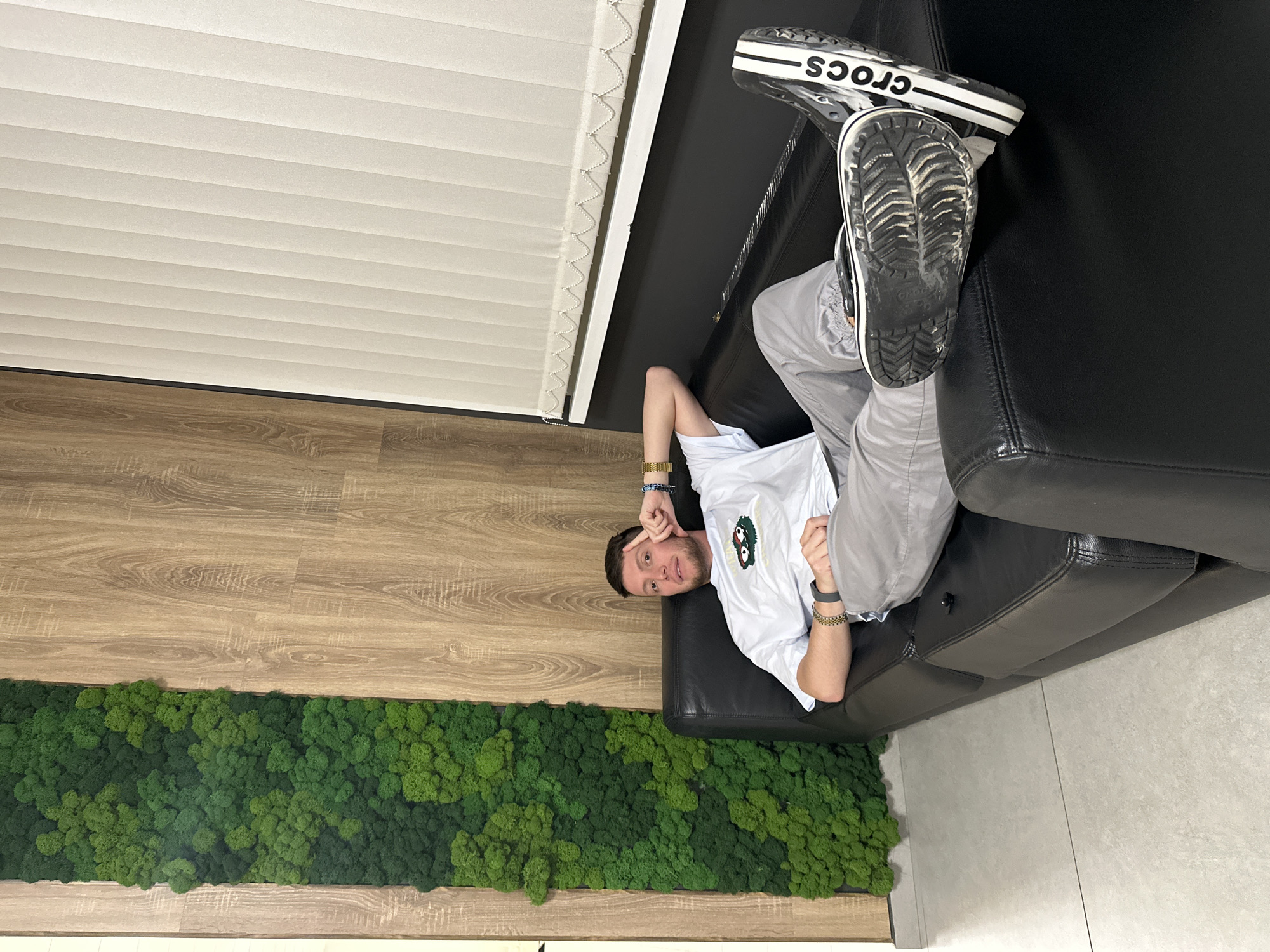 A man lies on a black leather sofa in what appears to be an office or modern living space. He is wearing grey pants, a white shirt with a small graphic, and black and white Crocs. He is lying with his head towards the viewer's left, and his feet are propped up on the back of the sofa. The room has a wooden floor, a green moss wall feature, and white window blinds. The location is not discernible as Moscow, Russia.