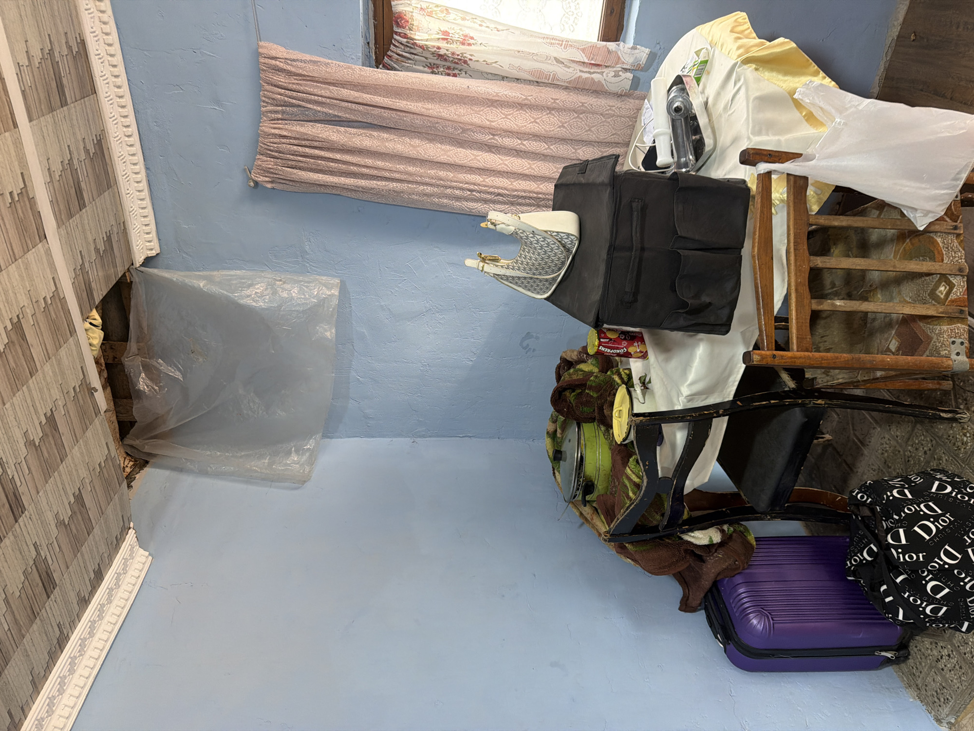 This is a photograph of an interior scene, likely a room in a house, located in Köhnə Quşçu, Azerbaijan. The room features pale blue walls and a patterned ceiling. A significant portion of the ceiling appears damaged, with exposed materials and a piece of plastic sheeting draped over the hole. A white decorative molding runs along the top of the walls, but it is detached and damaged near the ceiling opening.
In the center of the room, a round table is covered with a white and yellow tablecloth. On the table, there are several items: a large black bag with multiple compartments, a smaller white and gray handbag, a green pot with a lid, a brown blanket or throw, a yellow-lidded container, and a tin of what appears to be cookies. A metal tray with some items is also on the table.
Two chairs are positioned around the table. One chair, partially visible, has a black seat and a wooden frame. Another chair, made entirely of wood with a woven seat, is visible to the right. A purple suitcase stands on the floor to the left of the table, next to a black bag with a repeating "Dior" logo pattern. A white plastic bag is hanging from the wooden chair. A window with pink patterned curtains is visible on the right side of the image. The floor appears to be tiled. There are no people present in the scene.