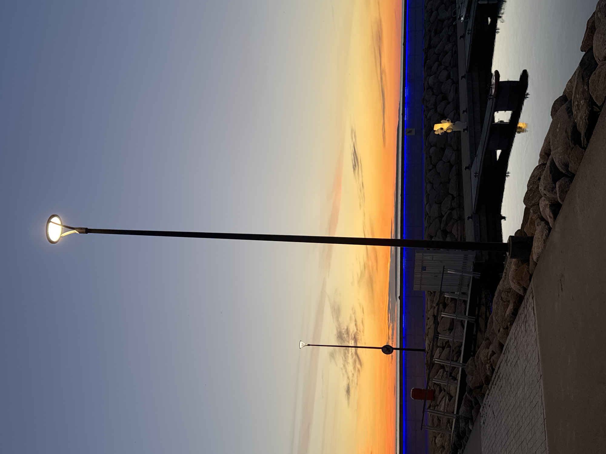 The image shows a harbor scene at dusk in Tallinn, Estonia, likely in the Pirita marina area, known for its modern piers and waterfront promenades. A tall, dark lamppost with a lit, circular fixture dominates the center-right of the frame, its light illuminating the sky. In the background, the sky transitions from a pale blue at the top to vibrant orange and yellow hues near the horizon, indicating sunset or sunrise. A thin, bright blue light runs horizontally along the waterfront barrier.
In the mid-ground, a paved walkway with cobblestone detailing runs along the left edge. To the right of the walkway, a rocky embankment leads down to a marina. Several boats are docked, with their reflections visible in the calm water. A small metal bridge extends from the walkway towards the boats. Another, smaller lamppost is visible on the walkway further in the distance, with a red cylindrical object nearby. The water is dark, reflecting the sky and the lights of the marina. The overall atmosphere is serene and tranquil.