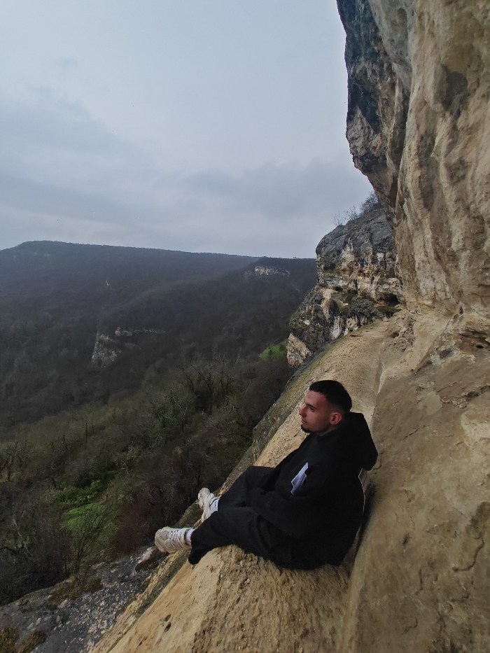 The image displays a male individual seated on a narrow, light-colored rock ledge overlooking a densely forested valley under an overcast sky. This scene is consistent with the rugged terrain found in the Shumen Plateau nature park, located near Shumen, Bulgaria. The subject, a young adult male with short, dark hair, is positioned in the lower right portion of the frame, facing right and looking out across the valley. He is wearing a black hooded sweatshirt with a white geometric logo on the left chest, black pants, and light-colored athletic shoes with white laces. His posture is relaxed, with legs extended slightly forward. The prominent cliff face, composed of stratified rock formations and minimal vegetation, dominates the right side of the image, while the vast expanse of the valley, covered in dark green deciduous and evergreen trees, recedes into the background under a uniformly grey, cloudy sky.