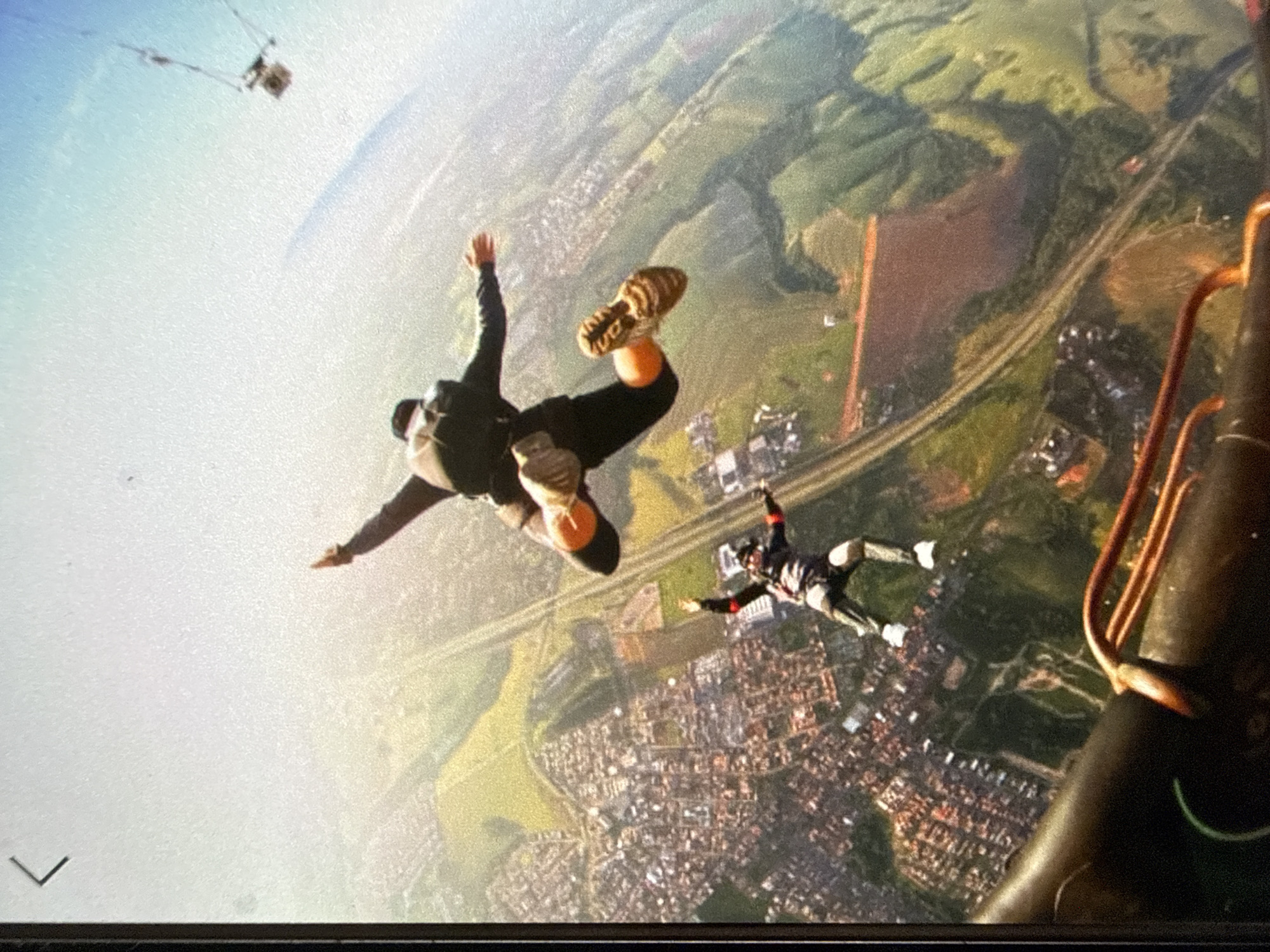 The image shows two skydivers in freefall over a city. One skydiver is oriented with their feet pointing upward, wearing dark clothing and tan-colored shoes. The other skydiver is oriented with their head pointing downward, wearing a patterned suit. The city below is a mix of buildings, roads, and green spaces. A road network is clearly visible. There are additional items in the air above. A section of an aircraft is visible on the right side of the image. The location and city are indeterminate.