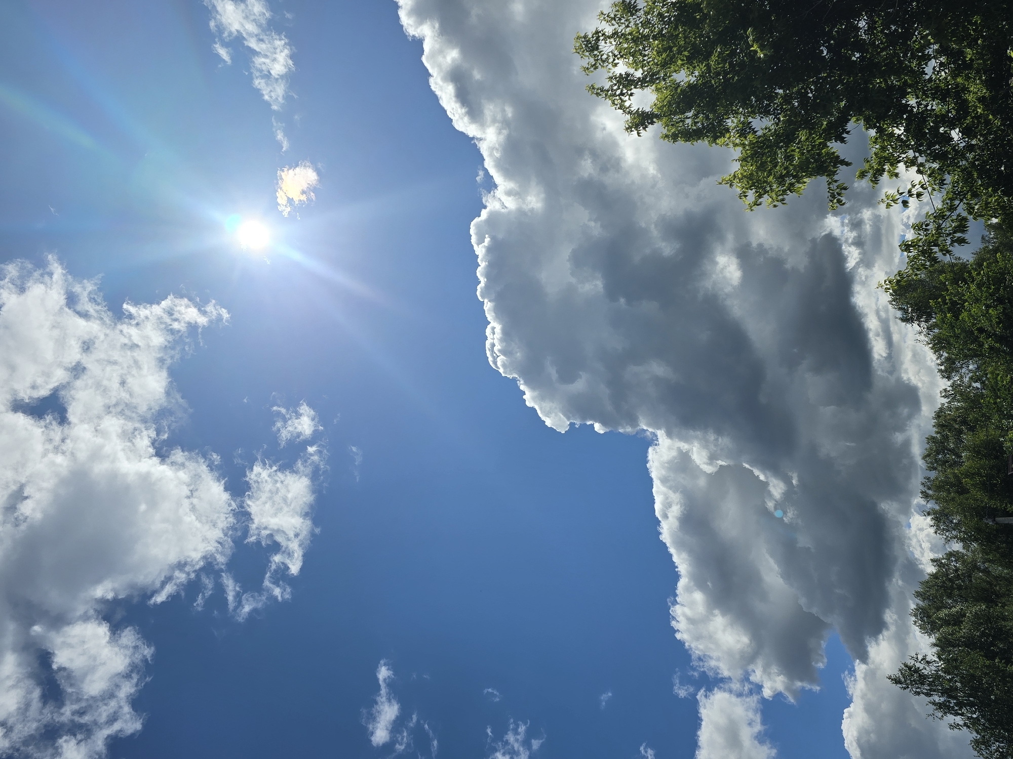 The image depicts a sky with a vibrant blue expanse, partially obscured by a mix of fluffy white cumulus clouds and darker, more ominous storm clouds. The sun is shining brightly, creating a lens flare effect and casting rays across the scene. In the lower portion of the image, a dense line of green trees forms a canopy, suggesting an outdoor environment. The overall impression is one of dynamic weather conditions, possibly in a natural or semi-urban setting. The location is identified as Ермолаево, Russia.