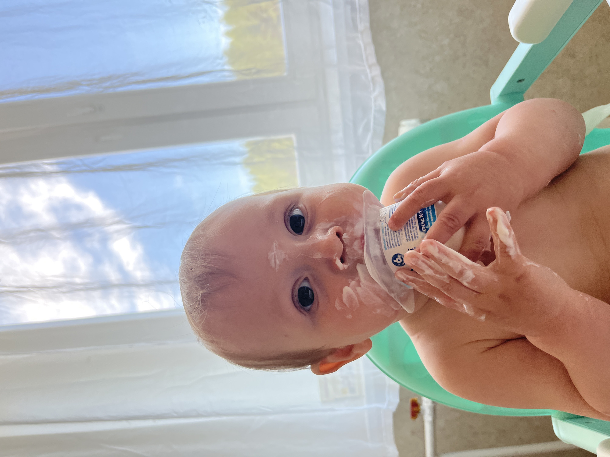 A baby is captured mid-activity, looking directly at the camera with wide, dark eyes. The baby's face, especially around the mouth and cheeks, along with both hands, is covered in a white, creamy substance, suggesting a messy eating or play session.
The baby has fair skin and light-colored hair. It is holding a white, squeezable pouch or container with both hands, with some of the white substance also on the pouch itself. Visible Cyrillic text on the pouch includes "БРЕБИ" (likely part of a brand name), "ЯГ" (possibly an abbreviation for "ягоды" - berries), and "No1", along with a blue and white circular logo featuring a number and an arrow.
The baby appears to be seated in a light green or turquoise high chair or baby seat. In the background, a large window reveals a bright blue sky with white clouds, indicating it is daytime. Parts of the window frame or blinds are also visible. The setting is indoors, likely in a home in Chaikovsky, Russia, where the image was taken. The overall scene conveys a playful and engaging moment, typical of a young child's mealtime or sensory exploration.