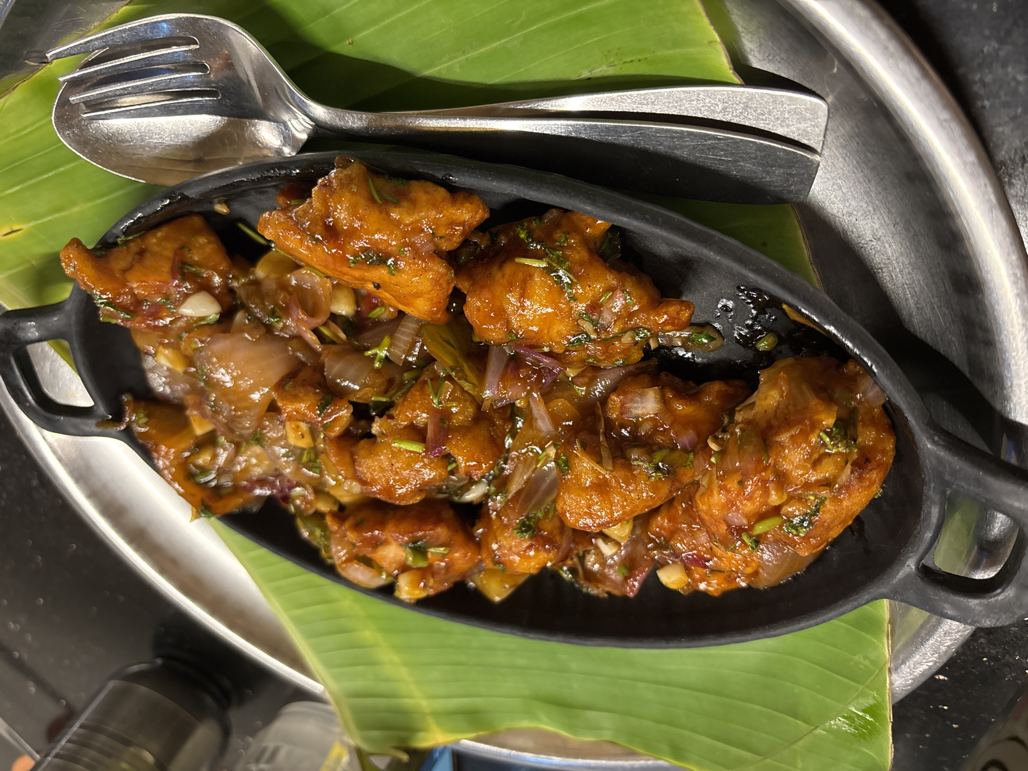The image depicts a close-up of a serving of Chicken Manchurian, a popular Indo-Chinese dish, presented in a black oval serving dish. The dish rests on a vibrant green banana leaf, which in turn lies on a larger, silver-colored platter. A serving spoon and fork are placed beside the serving dish on the banana leaf. The Chicken Manchurian appears to consist of bite-sized pieces of fried chicken coated in a dark brown, slightly glossy sauce. Diced onions and green herbs are visible within the sauce. The image was taken in Kamalapura, India.