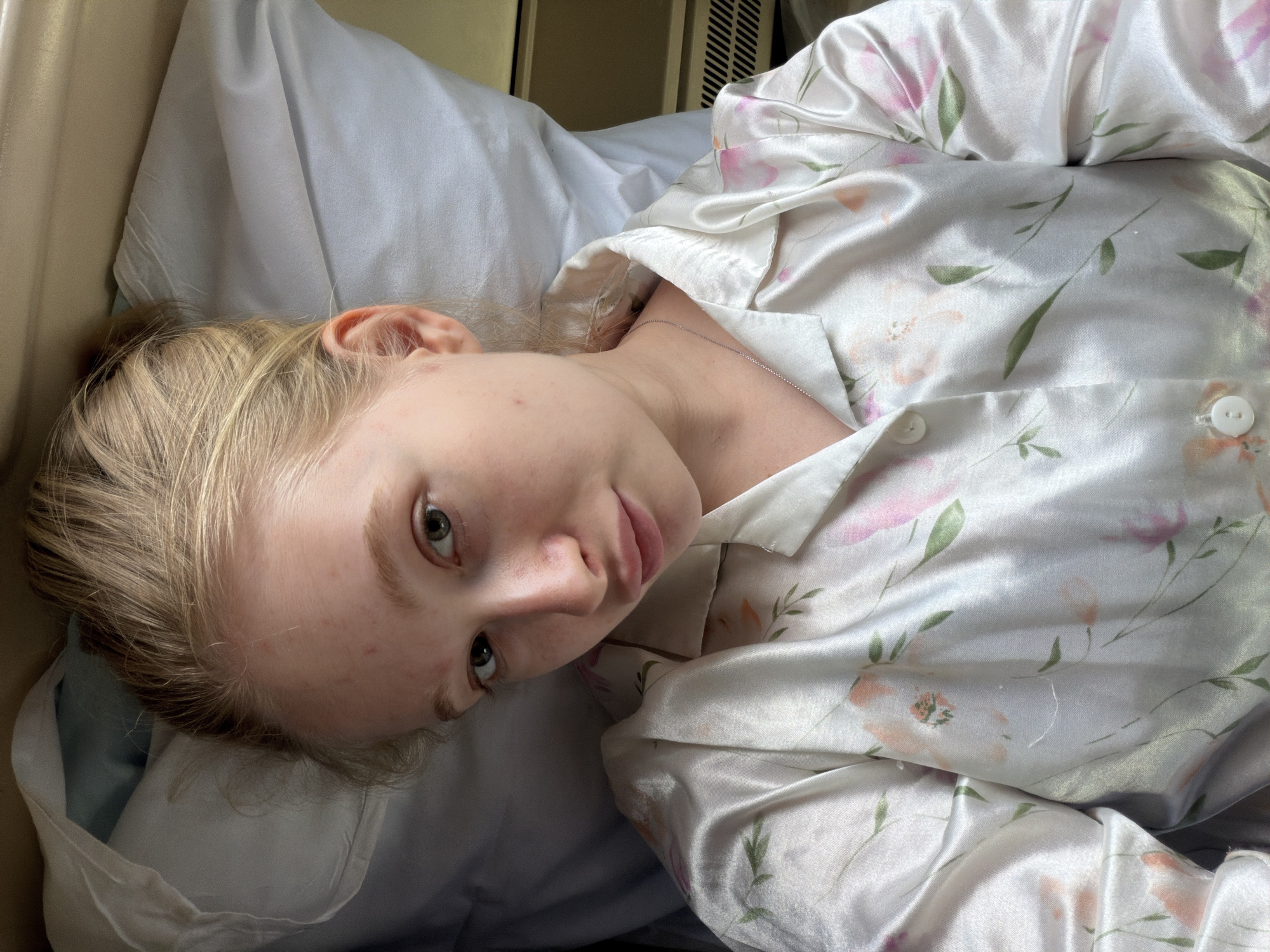 A fair-skinned person with blond hair is lying on a white pillow and appears to be in bed. They are wearing a satin pajama top with a floral pattern of peach-colored flowers with green leaves and pink accents. The background includes part of a white headboard or wall, and a beige wall or panel. The location is indiscernible from the image alone; therefore, Krasnodar, Russia is an unsupported assertion.
