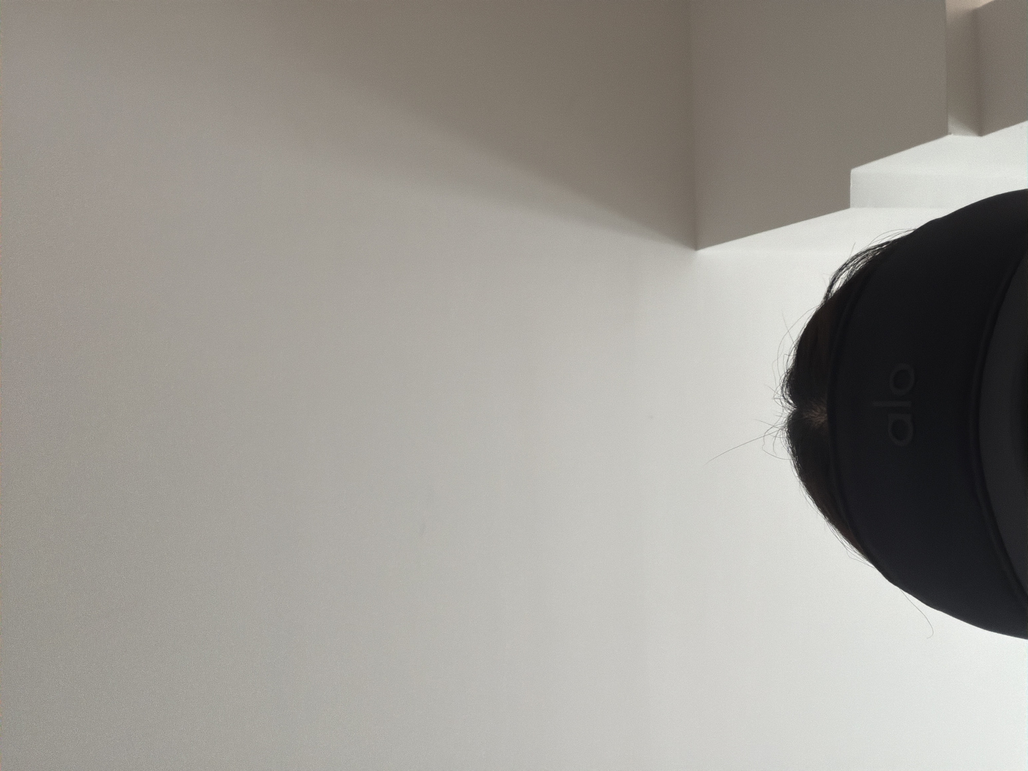 A close-up, slightly rotated image shows a person wearing a black "alo" branded visor. The background consists of a corner of a white wall and what appears to be a portion of a white shelf or molding. No definitive information allows pinpointing a specific location in Villahermosa, Mexico.