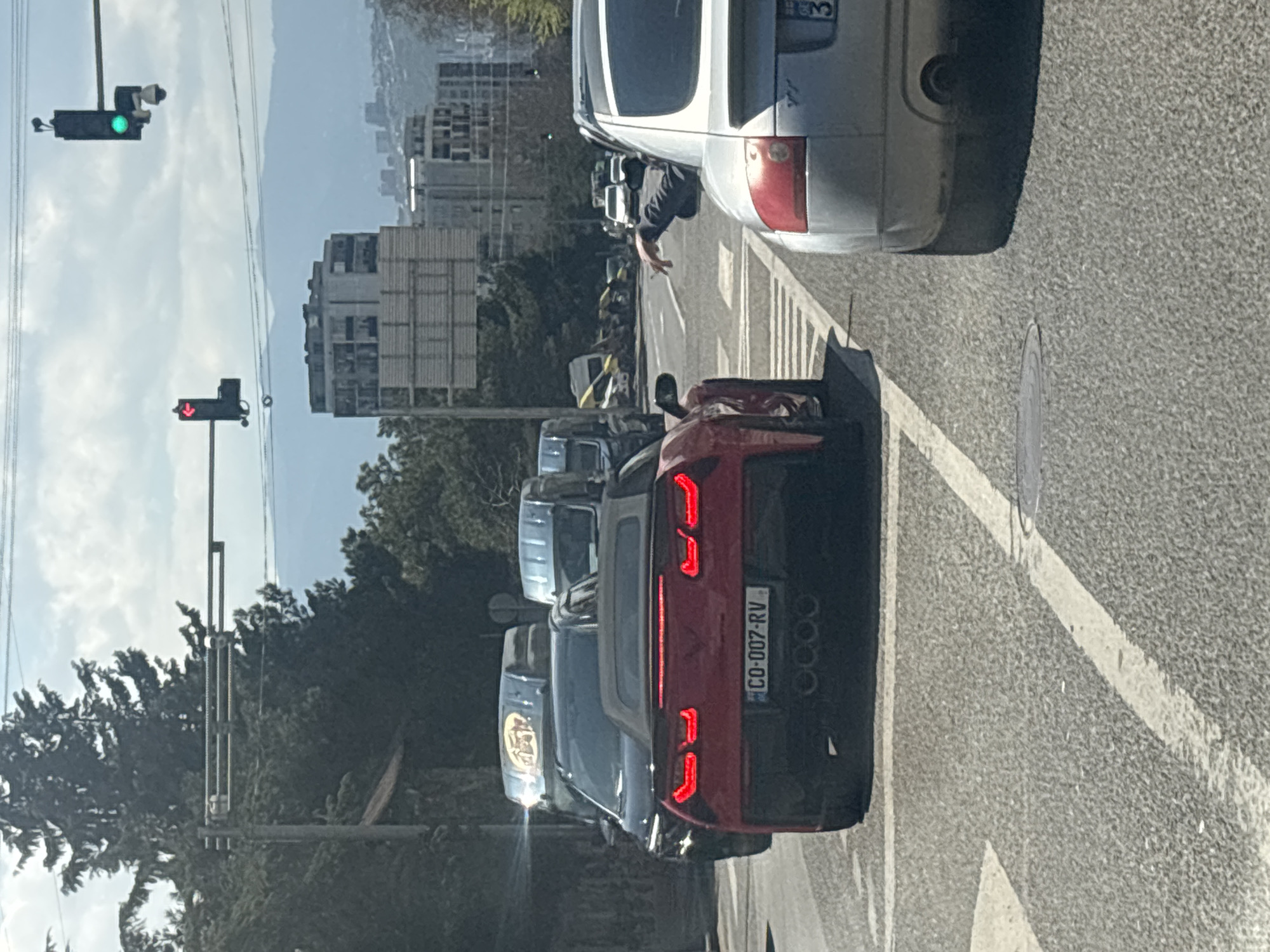 The image shows a street scene in Tbilisi, Georgia. The traffic is stopped at a red traffic light with a red arrow, while the pedestrian traffic light is green. A red car with license plate "CO-007-RV" is in the foreground. A person is seen reaching their hand out from the window of a silver car in the next lane. Other vehicles are visible in the background, along with buildings and trees. The weather appears sunny.