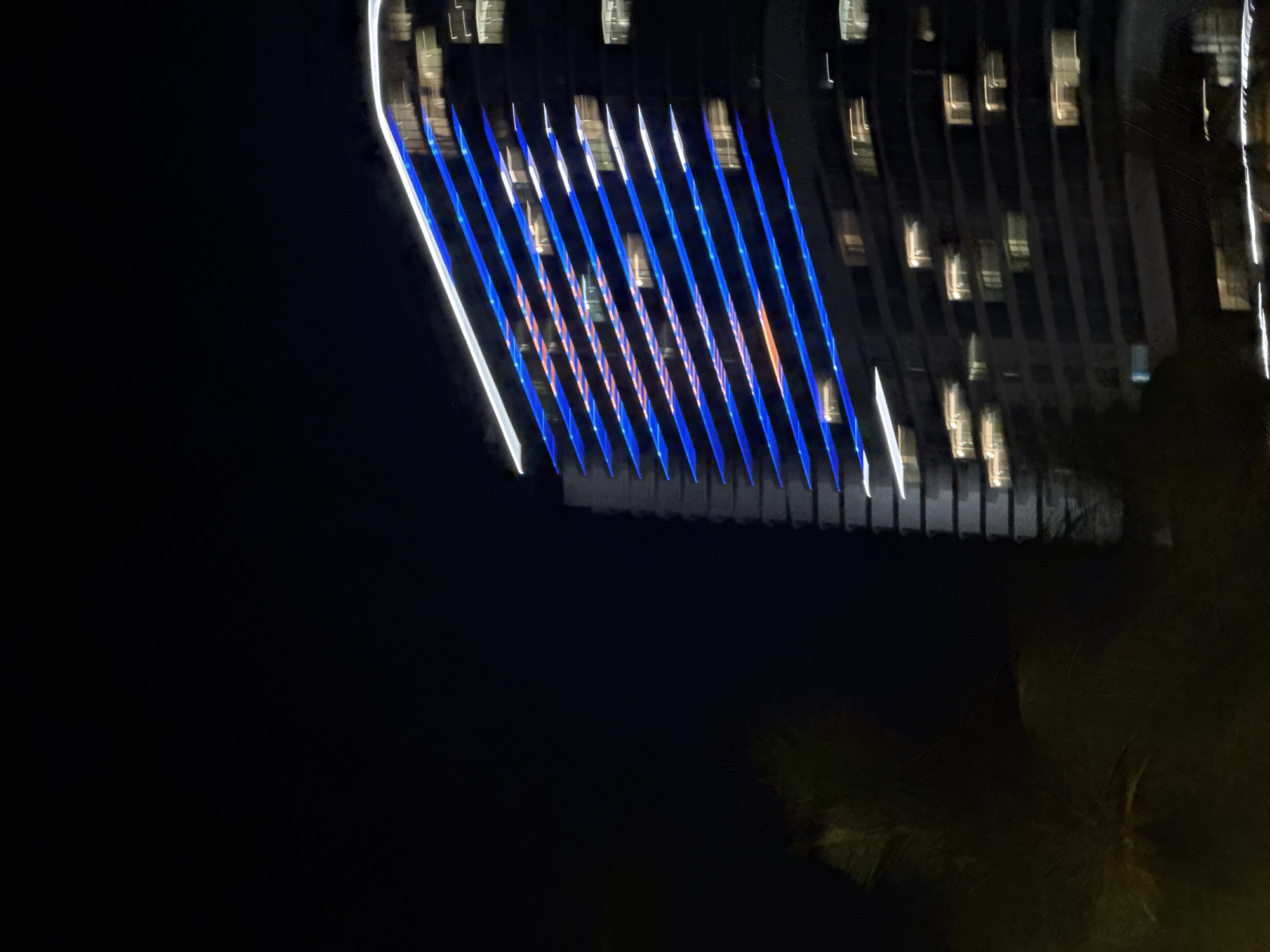 A blurry, nighttime photograph shows a tall building with diagonal blue and white lights along its exterior. The building appears to be made of dark material with many windows, some of which are illuminated with a yellowish light. The bottom left of the image shows out-of-focus green foliage, possibly from a tree or shrub. The overall impression is of a modern building at night, with its lighting system creating a striking visual effect against the dark sky. The location is identified as Bac Nha Trang Ward, Vietnam.