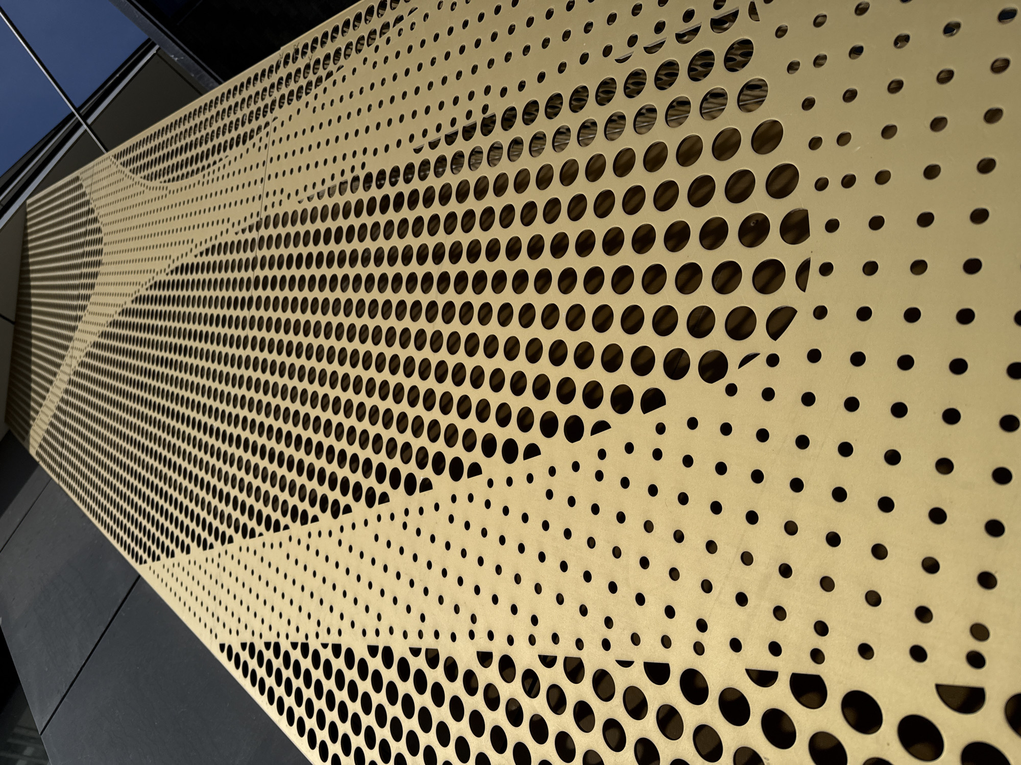A tan metal panel with circular cutouts of varying sizes covers a portion of a building in Yekaterinburg, Russia. The panels are angled upwards and to the left, creating a perspective view. The background shows sections of dark gray building materials and a glimpse of blue sky. The circular cutouts create a patterned effect on the tan metal.