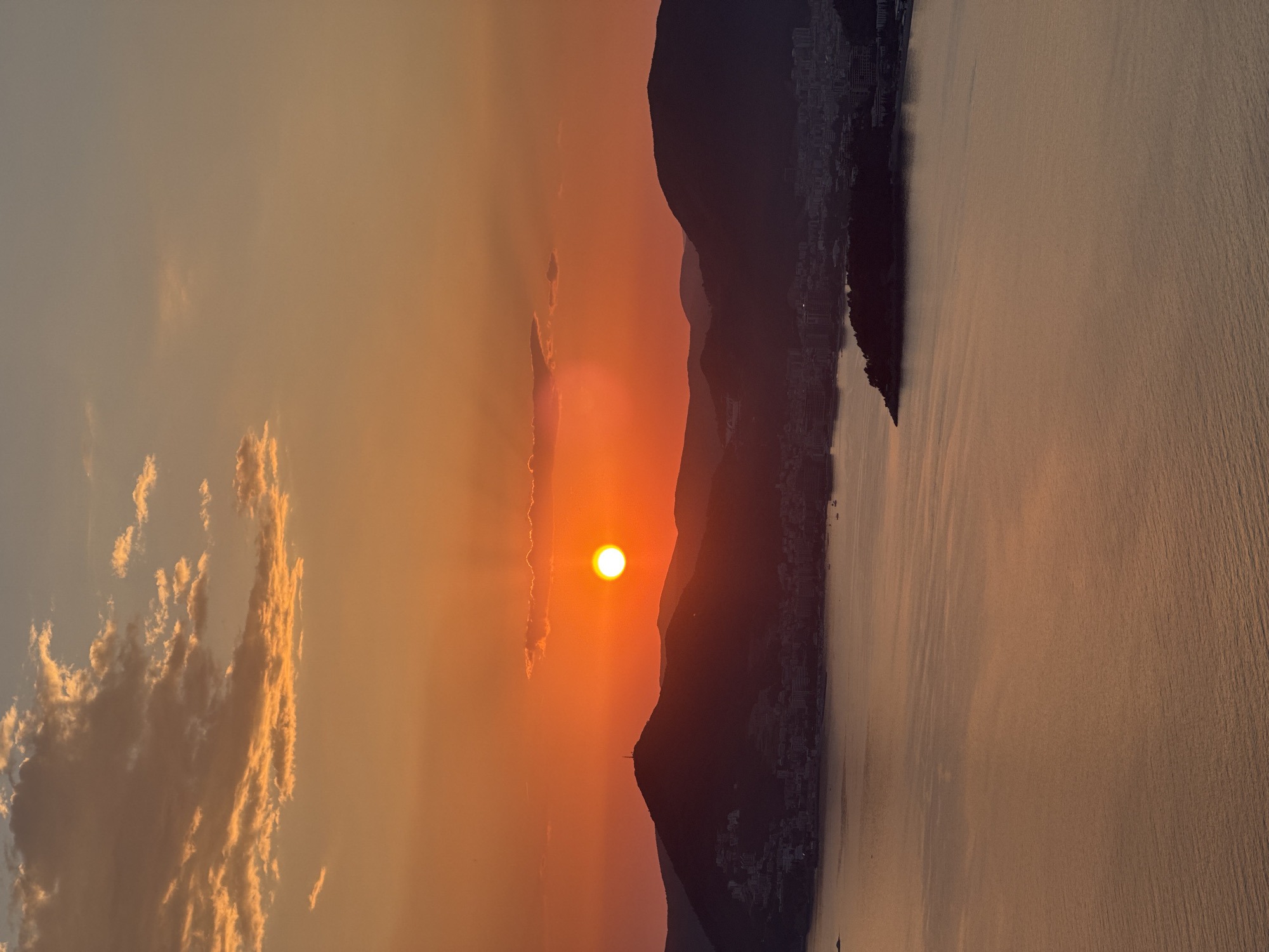 The image shows a sunset in Sveti Stefan, Montenegro. The sun is positioned in the center of the frame, casting an orange glow across the sky and water. The silhouette of a mountain range is visible between the sun and the water. The dark shape of the island of Sveti Stefan is at the right. Clouds are visible in the sky on the left. The sea reflects the colors of the sunset.