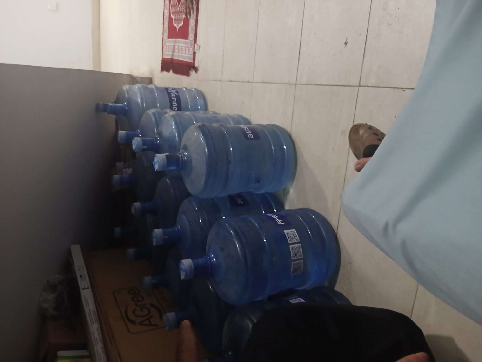 In the city of Peshawar Tehsil, Pakistan, a dimly lit room contains several large, blue plastic water jugs arranged on a tiled floor. Some jugs have white caps and visible labels with QR codes. In the background, a brown cardboard box with the "AGree" logo is partially visible. To the right, a red prayer rug is laid out on the floor, and a bare foot wearing a brown sandal is positioned near the rug. The overall environment suggests a storage area or a room where water supplies are kept.