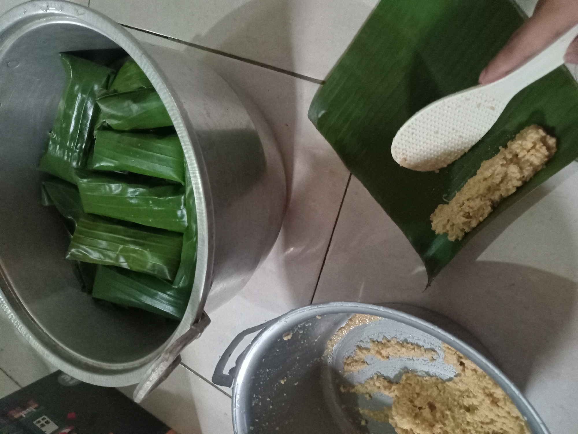 In Claket, Indonesia, this image captures a close-up view of traditional Indonesian food preparation. Two aluminum pots are visible on a tiled floor. One pot, larger and situated in the upper portion of the image, contains several food items wrapped in green banana leaves, neatly arranged inside. The other, smaller pot, located in the lower-left quadrant, holds a portion of the light brown, granular food mixture, possibly rice or a similar grain-based preparation. To the right of the smaller pot, a banana leaf is laid out, upon which a line of the same food mixture has been placed. A white rice paddle is positioned next to the food, its textured surface evident. A person's finger is lightly touching the paddle, suggesting an action in progress. The background is indistinct, but the overall setting appears to be a domestic environment, likely a kitchen.