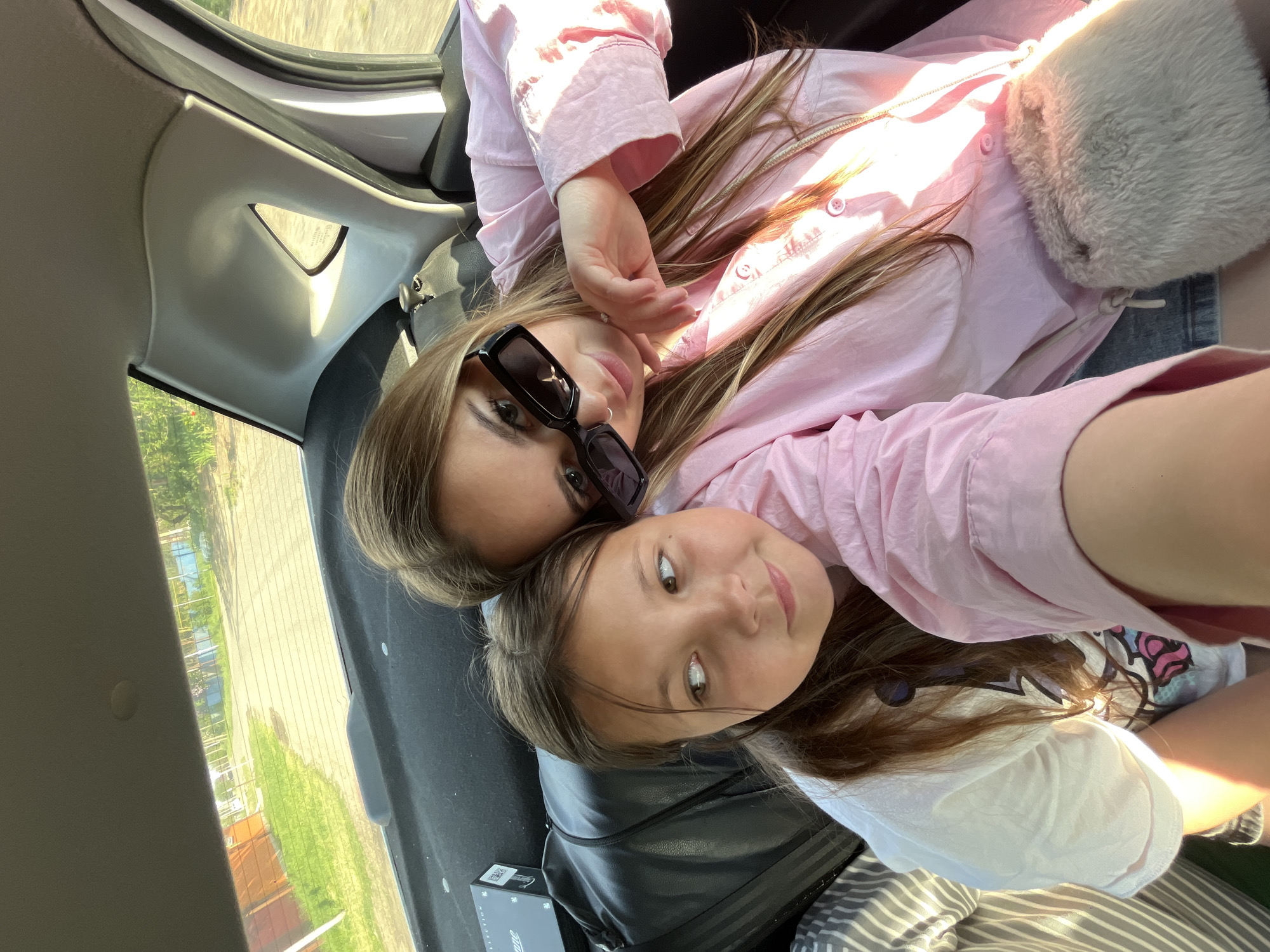 Two females are lying down inside the trunk of a car, angled towards the camera. The older female is wearing sunglasses and a light pink, button-down shirt. The younger female is wearing a white t-shirt. Outside the car, a rural area with green grass and trees can be seen. The location is Novopokrovskaya, Russia.