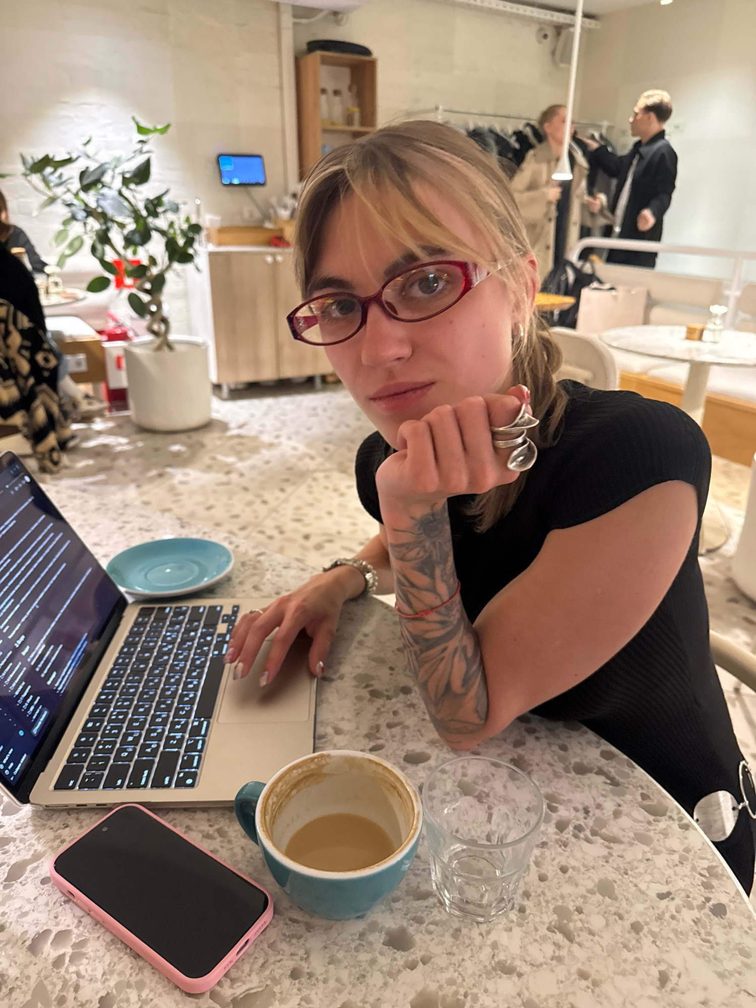 A young woman with blonde hair and glasses is sitting at a table in a brightly lit, modern cafe, likely in Moscow, Russia. She is looking at the camera, with her right hand resting on her chin, and her left hand poised over a laptop. Her left arm, visible in the foreground, is covered in a large tattoo. On the table in front of her are her open laptop, a pink-cased smartphone, a light blue teacup with coffee and foam, a small blue saucer, and a clear glass with ice. In the background, other cafe patrons can be seen at tables and a rack of clothing is visible. The cafe has a terrazzo-style flooring and light-colored walls. A potted plant with dark green leaves is situated to the left of the woman.