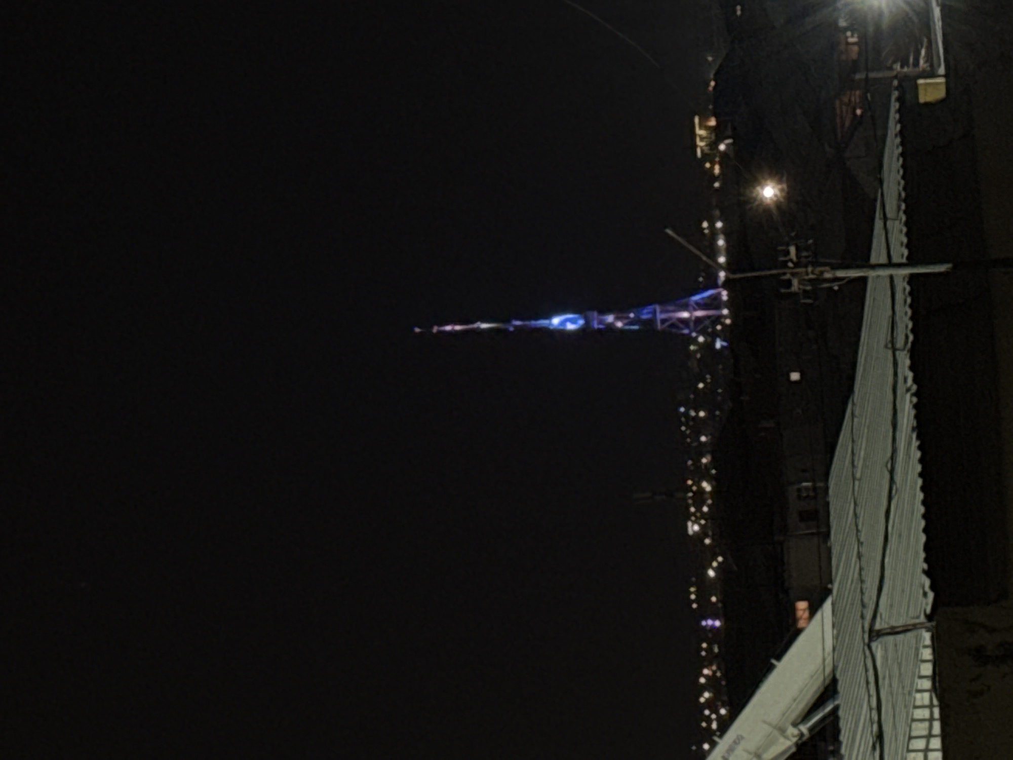The image captures a night scene in an urban environment, with a tall, illuminated tower as the central distant subject. The tower, likely the Yerevan TV Tower given the location context, is slender and emits distinct blue and purple light from its upper sections, contrasting against the dark night sky.
In the foreground, on the right side of the frame, is a close-up section of a building or structure. It features a prominent corrugated metal facade and is dimly lit by scattered light sources. Various bright, star-like lights are visible across the scene, suggesting distant city lights or other artificial illumination in the cityscape. A bright, lens-flared light source is situated near the top right, and another fainter one is in the mid-right. Wires or cables are discernible stretching across parts of the mid-right.
At the bottom right, a segment of a white, elongated structure, possibly a crane arm or a part of a building's exterior machinery, is visible. There appears to be faint, dark text on this white structure, but it is largely illegible. The scene is static, depicting the city after dark under a clear sky.