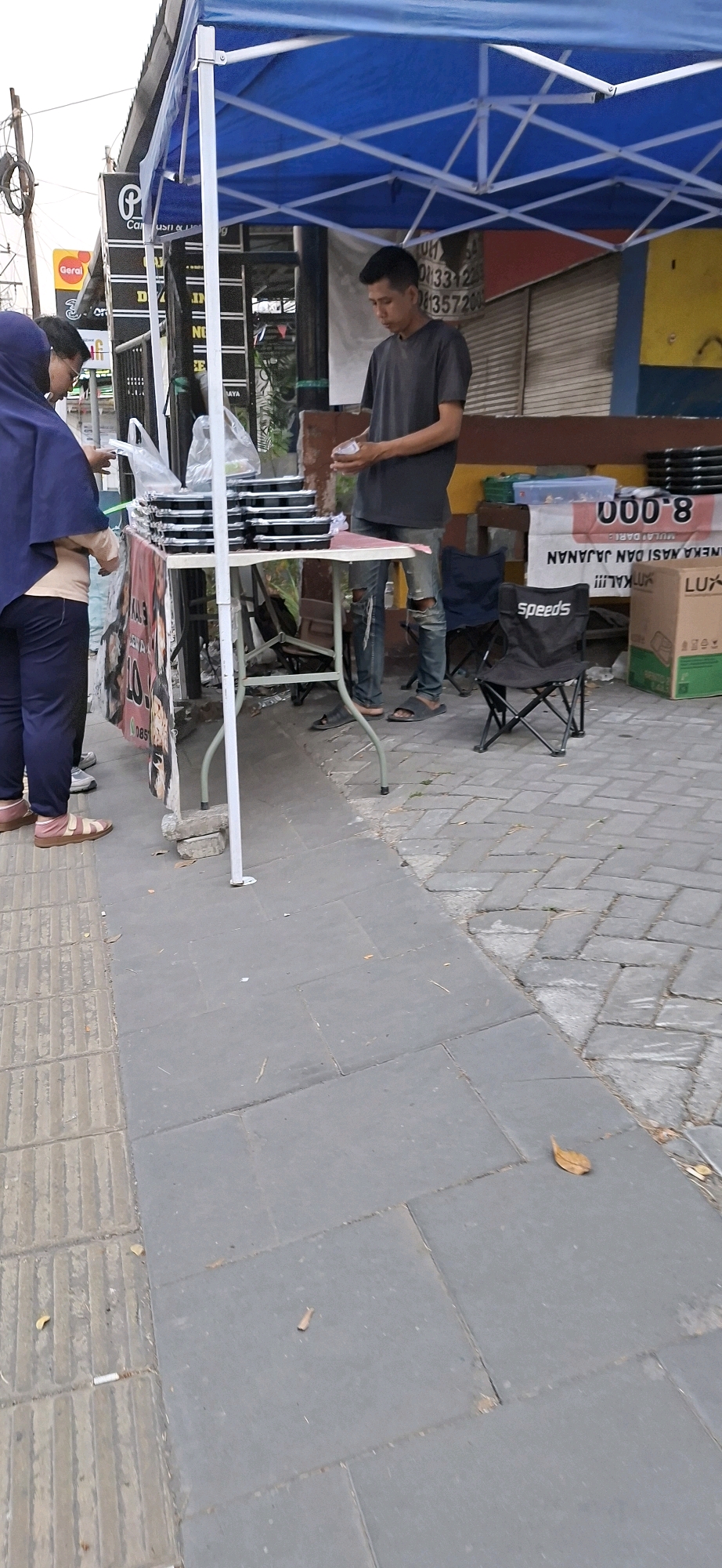 A low-angle, full-shot photograph captures a street vendor in Surabaya, Indonesia, under a blue canopy. A male vendor, wearing a dark grey t-shirt and ripped blue jeans, stands behind a folding table laden with pre-packaged meals in black containers. He holds a few small items, possibly cash or small food portions, in his hands. To the left, a woman wearing a dark purple hijab and navy blue pants stands partially in frame, facing the vendor and holding a white plastic bag. The table is draped with a banner featuring Indonesian text and a phone number. Beside the table are several folding chairs, one black chair with the "Speeds" logo, and a large cardboard box labeled "LUX". The pavement is composed of gray square tiles and textured gray tiles. In the background, the facade of a building with yellow and blue sections is visible, along with signage indicating a business named "Geral". The scene suggests a commercial transaction taking place on a sidewalk in a city environment.