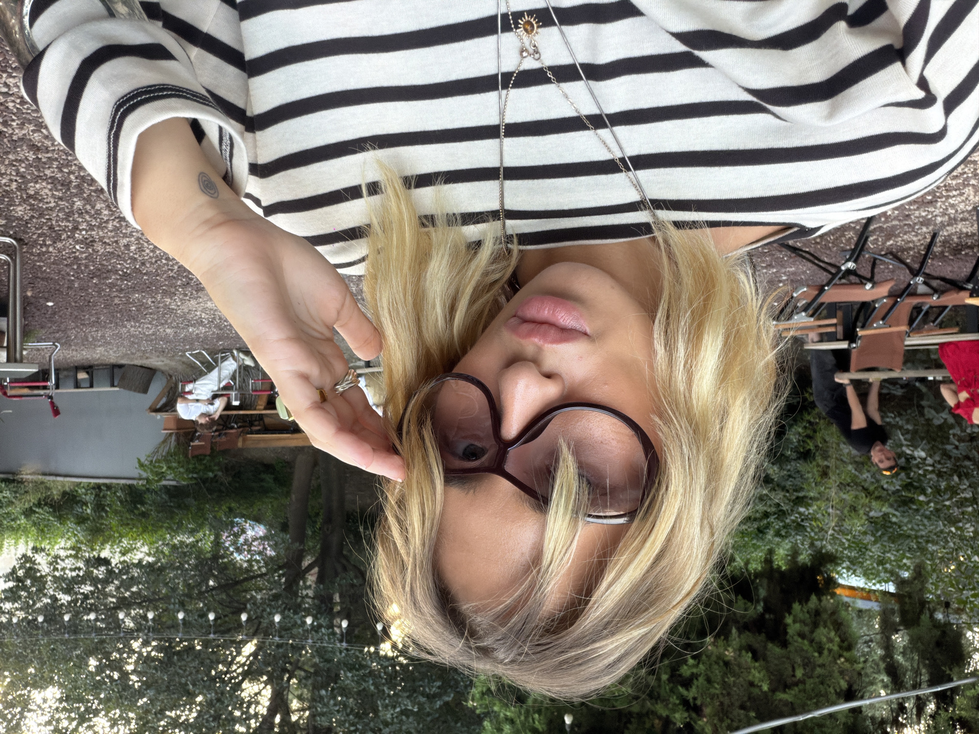 The image is a close-up, selfie-style portrait of a woman with blonde hair and large, dark-rimmed sunglasses. She is wearing a black and white striped long-sleeved shirt and multiple silver necklaces, one with a sun-shaped pendant. Her left hand is raised to her face, touching her sunglasses. A small, circular tattoo is visible on her inner wrist.
The background suggests an outdoor, possibly casual dining or social area. There are trees with string lights overhead, indicating it might be evening or dusk. In the middle ground, a man in a black t-shirt is visible, leaning on a table. Further back, another person is seated at a table. There are also outdoor chairs and tables scattered around. The ground appears to be covered in gravel or small stones. The overall ambiance suggests a relaxed, outdoor setting, potentially a cafe, restaurant, or garden area in Tbilisi, Georgia, given the prompt's context.