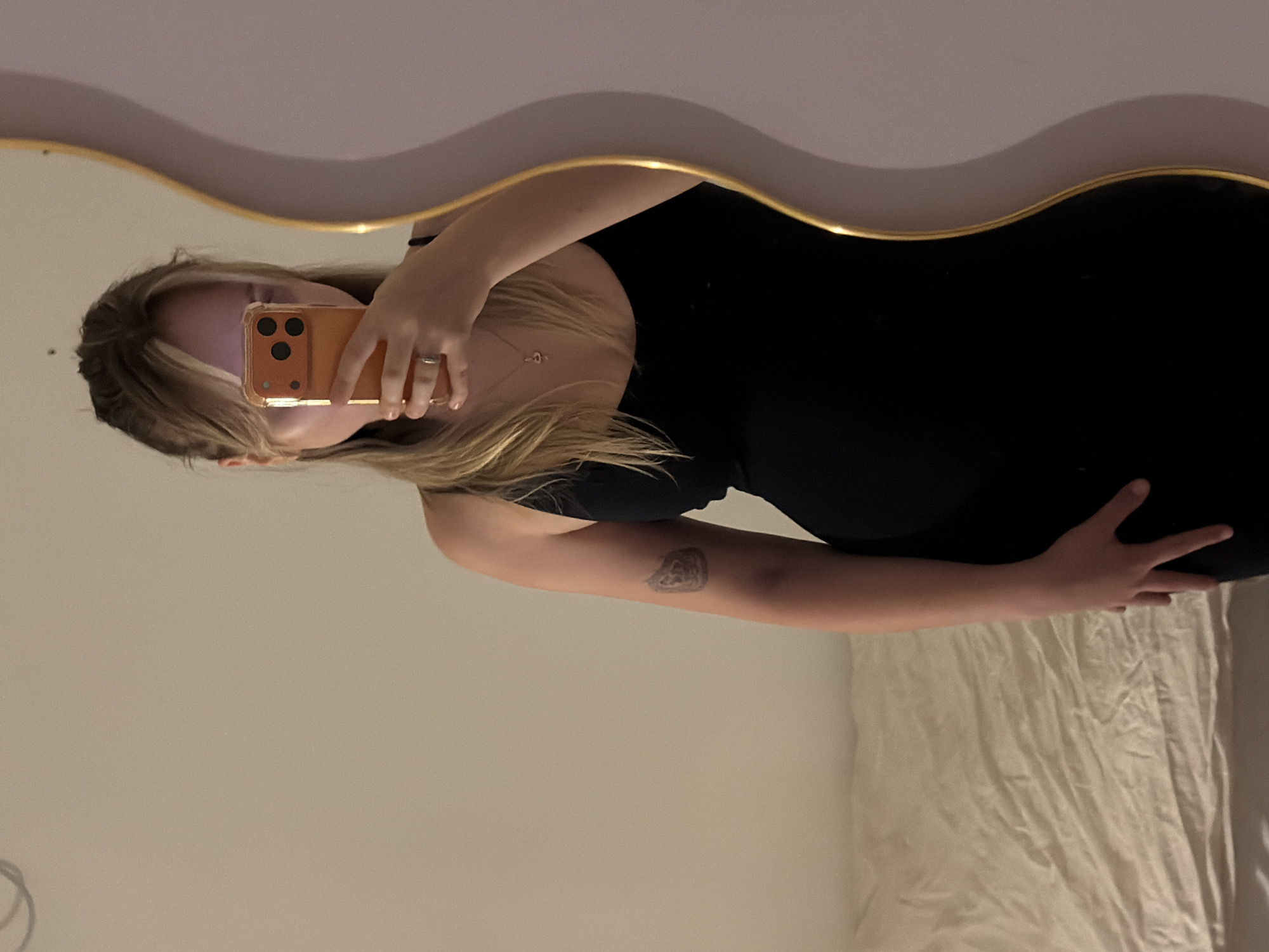 A fair-skinned person stands in front of a mirror with a gold frame against a light-colored wall, holding a mobile phone in their left hand and using it to take a selfie. The person is wearing a black dress and placing their right hand on their stomach. A small tattoo is visible on the person's left arm. Light-colored bed sheets are partially visible in the lower right corner of the frame. The location is not discernible from the image.