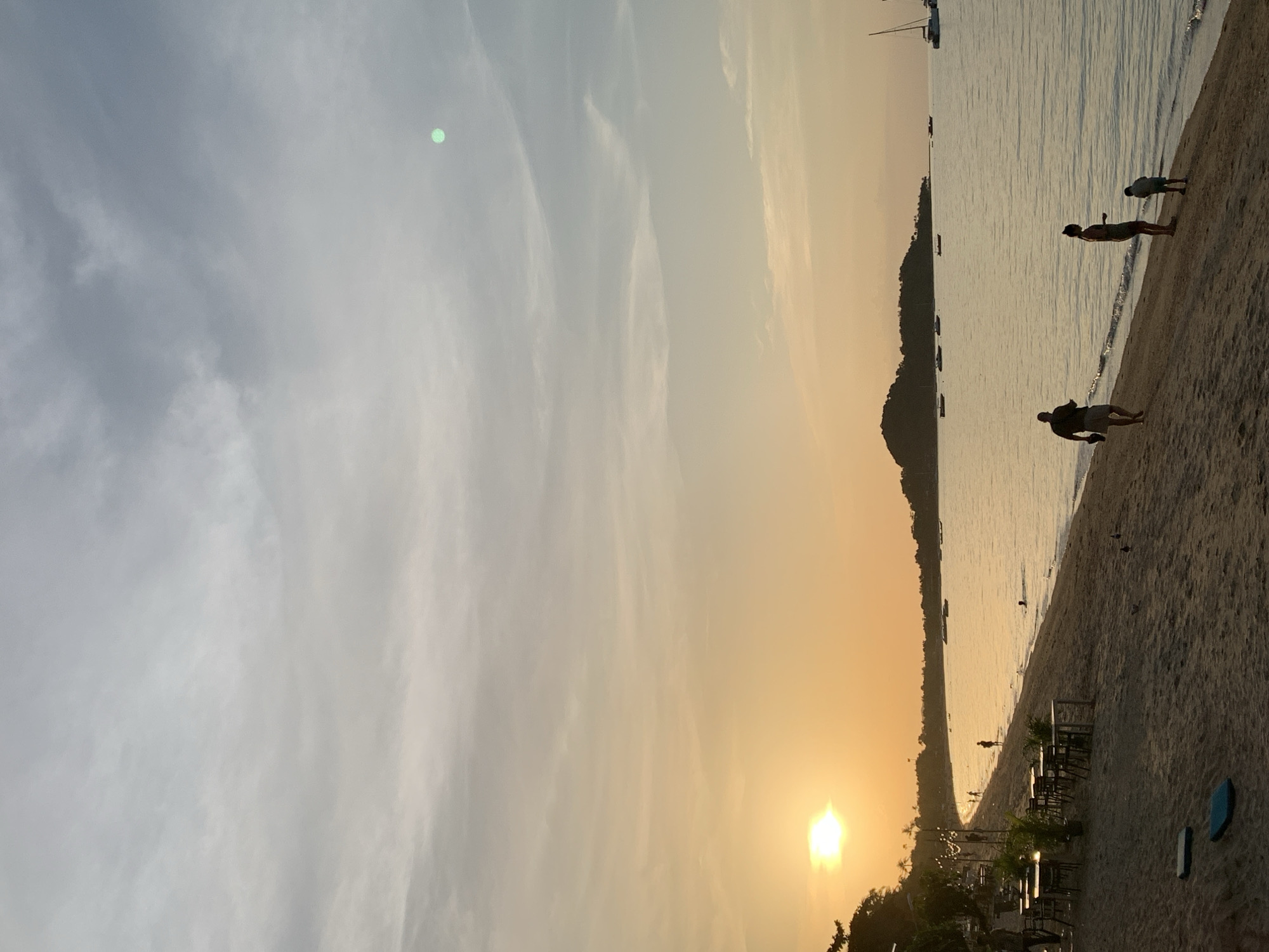 This is a vertical, medium-shot, eye-level photograph taken on a beach in the Ko Samui District of Thailand, likely during sunset. The sun, a bright yellow orb with a hazy halo, is low on the horizon to the left of the frame, casting a warm, orange glow across the sky and water. The sky is a gradient of soft oranges and yellows near the horizon, transitioning to pale blue and gray with scattered clouds higher up. A small, indistinct green circular object is visible in the upper right quadrant of the sky.
The foreground consists of a sandy beach with footprints and a few scattered blue mats or cushions. To the left, along the edge of the beach, are several sets of outdoor tables and chairs, some with potted plants, suggesting a beachfront dining area. String lights are visible above this area.
In the middle ground, the gentle waves of the ocean lap the shore. Several small boats and yachts are dotted across the water. In the distance, a silhouetted, hilly island rises from the sea.
Several people are present on the beach and in the water. On the sand, a man with his back to the camera is walking towards the water, carrying something in his right hand. Further down the beach, a woman and a young child stand near the water's edge, facing the sea. Another person is visible swimming or wading in the water. Other figures, appearing as small silhouettes, are scattered further along the beach and in the water. The overall atmosphere is serene and tranquil, characteristic of a tropical beach at dusk.