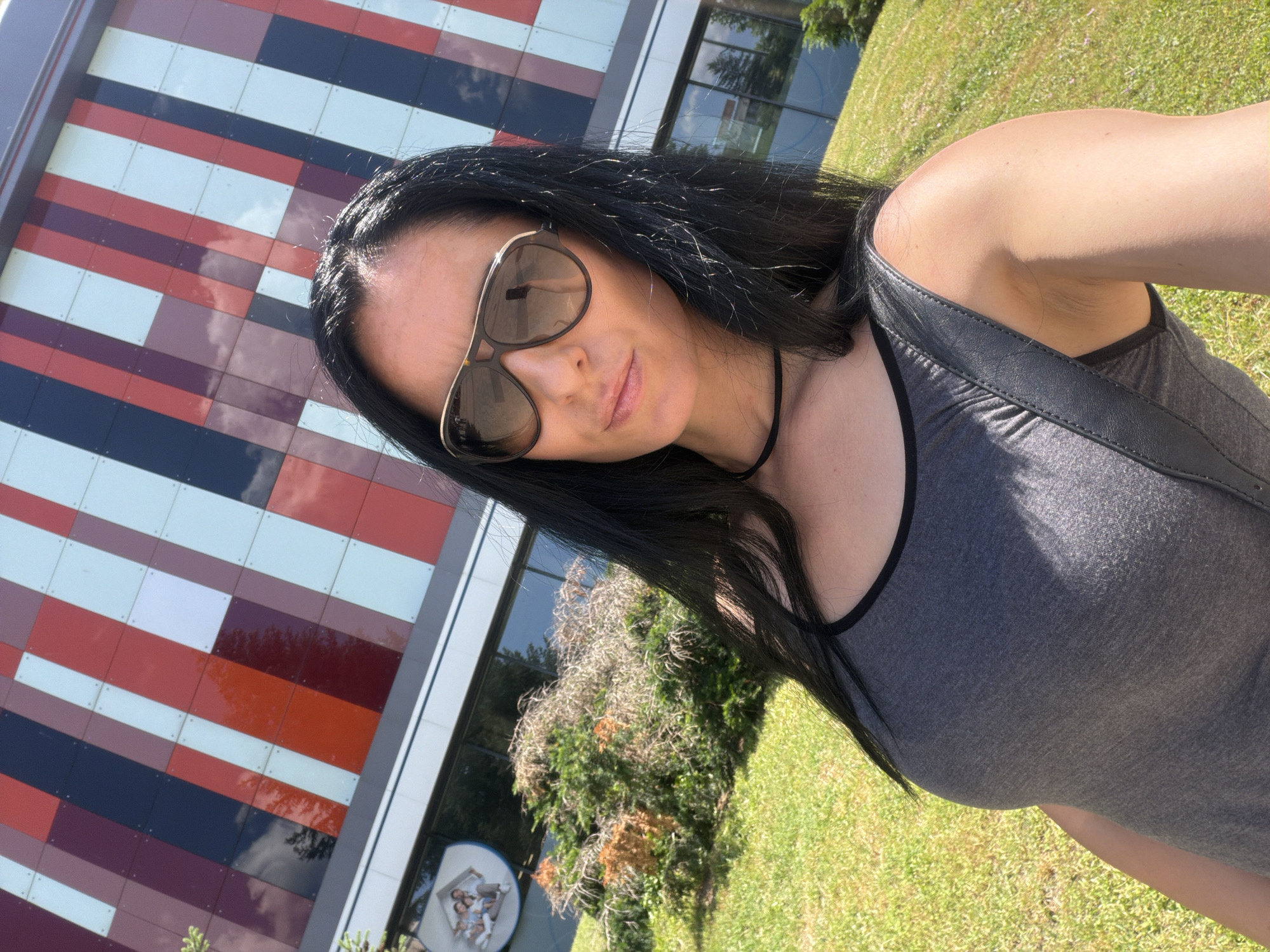 A woman is taking a selfie outside a modern building with a colorful, striped facade. She has long, dark hair and is wearing sunglasses and a gray tank top. The building appears to be a commercial or public establishment, possibly a cultural center or office building, given the signage visible on its exterior. The environment is sunny with green grass and some landscaping. The building's architecture suggests a contemporary design, and the colorful panels might be a distinctive feature of its facade. The image seems to be taken in an urban or suburban setting. While no specific landmark is explicitly identified as being in Pleven, Bulgaria, the query suggests this context.