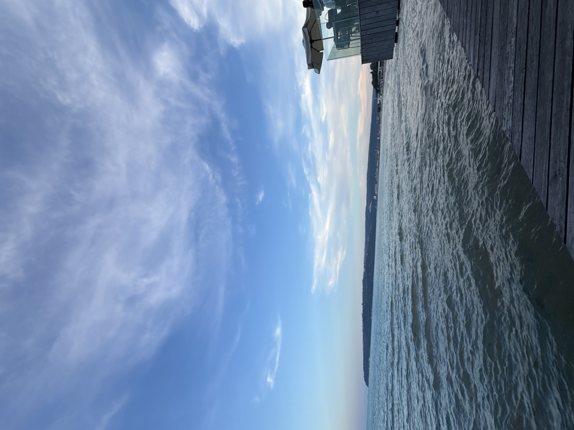 This image depicts a view of the sea and the Varna coastline from a wooden pier or deck. In the foreground, a weathered wooden deck runs diagonally across the bottom right of the frame, leading towards the water. The sea is choppy with small waves and appears to be a dark, greenish-blue color.
On the right side of the image, a modern outdoor seating area is visible, featuring glass railings, tables, and chairs. An umbrella shades a table, and two people are seated there, facing away from the camera.
In the distance, a long, hilly coastline stretches across the horizon, lined with trees and buildings, suggesting the urban landscape of Varna. The sky above is a clear blue with scattered, wispy white clouds, indicating fair weather. The overall impression is of a serene, seaside setting, possibly at a restaurant or cafe overlooking the Black Sea.