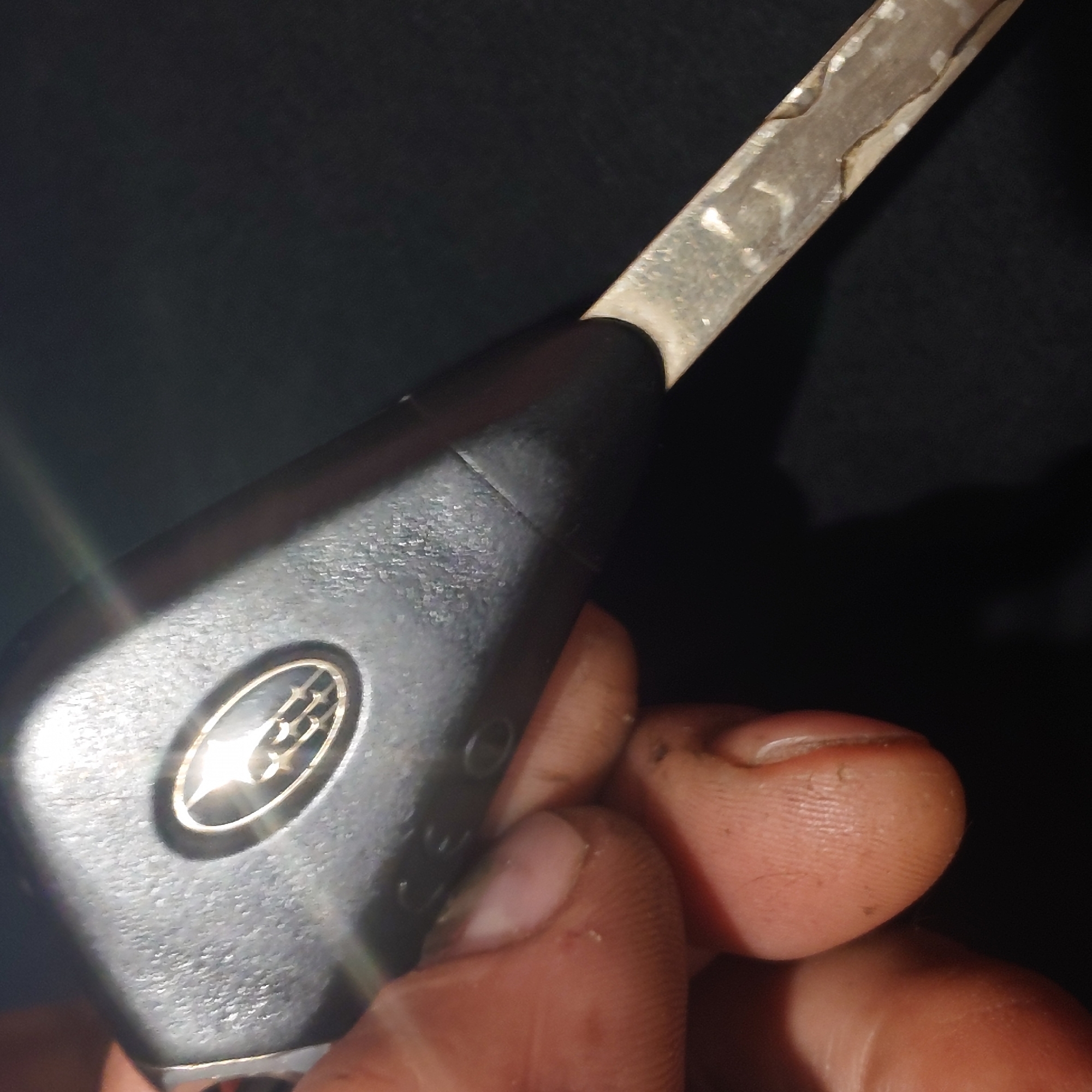 A close-up, low-angle shot shows a person's hand holding a Subaru car key. The key fob is black with a silver Subaru logo. The metal key blade is extended and appears to be worn. The background is dark and out of focus, with some light reflections visible on the key fob. No specific location in Belorechensk, Russia, can be identified from the image. The image is a still photograph.