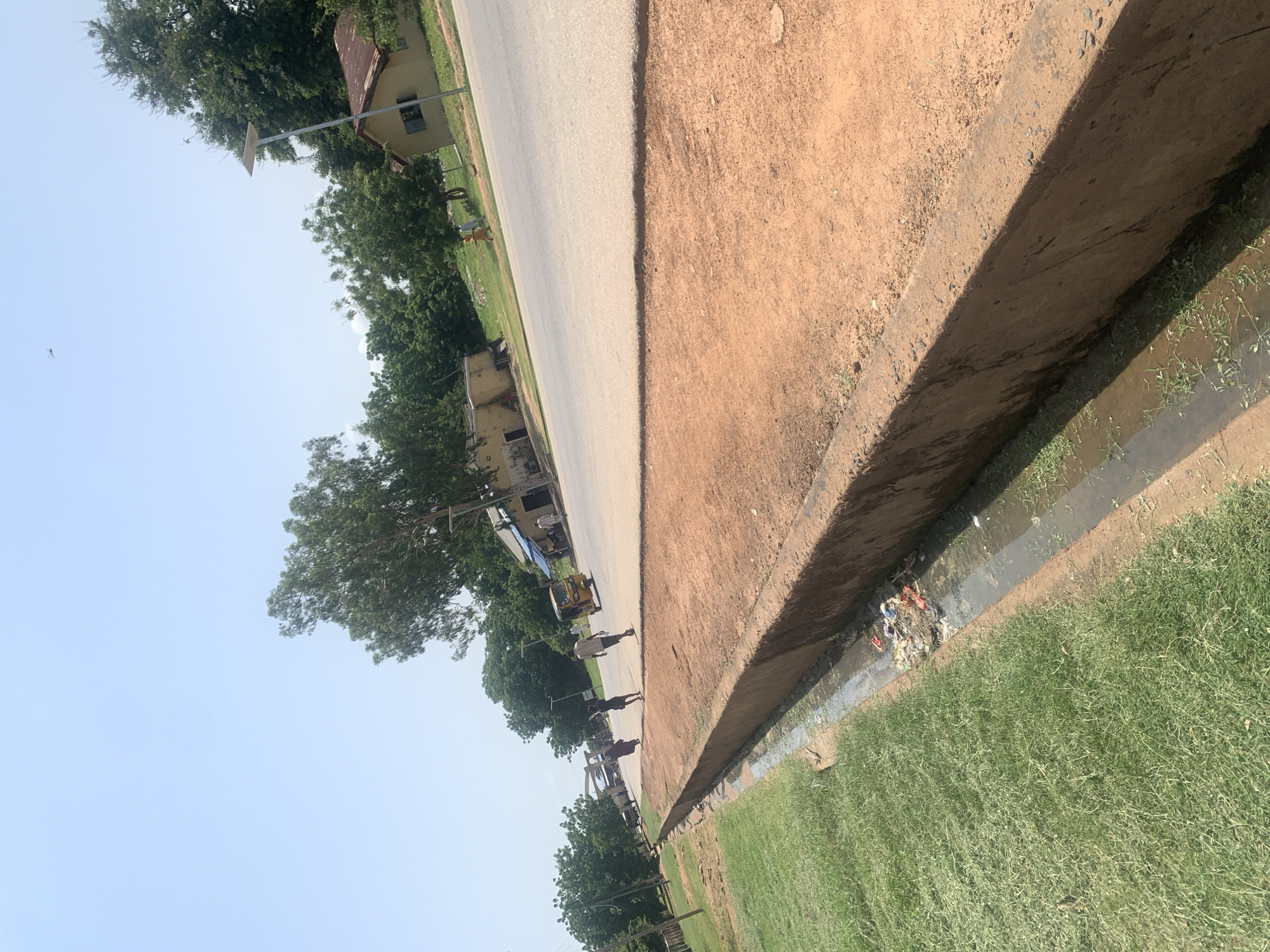 This image depicts a street scene in Jalingo, Nigeria. In the foreground, a grassy area is bordered by a concrete-lined drainage ditch filled with murky water and accumulated debris, including plastic bags and other refuse. The ditch is surrounded by reddish-brown earth. Beyond the ditch, a paved road runs diagonally through the frame, with a reddish-brown shoulder.
On the far side of the road, several buildings are visible, including a small, light-colored house with a corrugated iron roof and a larger structure with a blue awning. Trees and vegetation line the area behind the buildings.
Several people are present. Three individuals are walking away from the viewer on the road. One person is walking on the grassy area near the buildings. A yellow auto-rickshaw, commonly known as a Keke Napep, is parked on the road near some buildings, and another vehicle is further down the road. The sky is clear and bright, indicating a sunny day. In the sky, a single insect or drone is visible.
