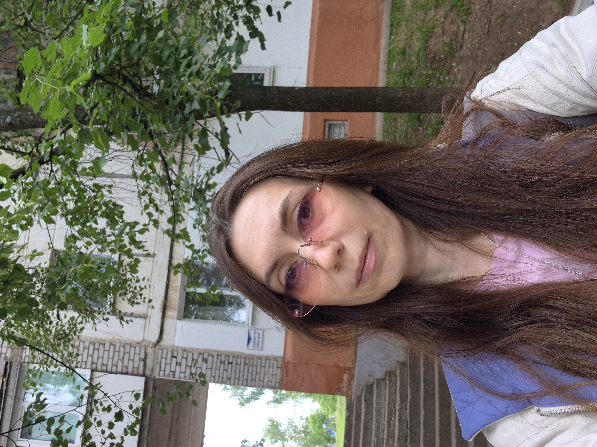 The image presents a young woman with long, dark brown hair, captured in an outdoor portrait with a tilted perspective. She is wearing rimless sunglasses with pink-tinted lenses, a pink top, a light blue jacket, and a white outer garment. A delicate silver chain necklace is visible around her neck. Her expression is calm and neutral, with a subtle hint of a smile, suggesting she is posing for a selfie.
The background features lush green foliage from several trees, indicating a spring or summer setting. Behind the trees, a multi-story building is visible, characterized by white walls, an orange-brown section, and a grey brick base. Numerous windows are embedded in the building's facade. To the right of the frame, a set of grey concrete or stone steps can be seen. The lighting is soft and diffused, characteristic of an overcast day or a shaded area during daytime.
A notable detail is a blue sign attached to the brick base of the building, displaying Russian text. The inverted text, when correctly oriented, reads: "ОБЪЕКТ ОХРАНЯЕТСЯ ВИДЕОНАБЛЮДЕНИЕМ," which translates to "OBJECT IS PROTECTED BY VIDEO SURVEILLANCE." This sign provides a strong location cue, aligning with the context of Chaikovsky, Russia, and placing the scene in an urban or suburban Russian environment.