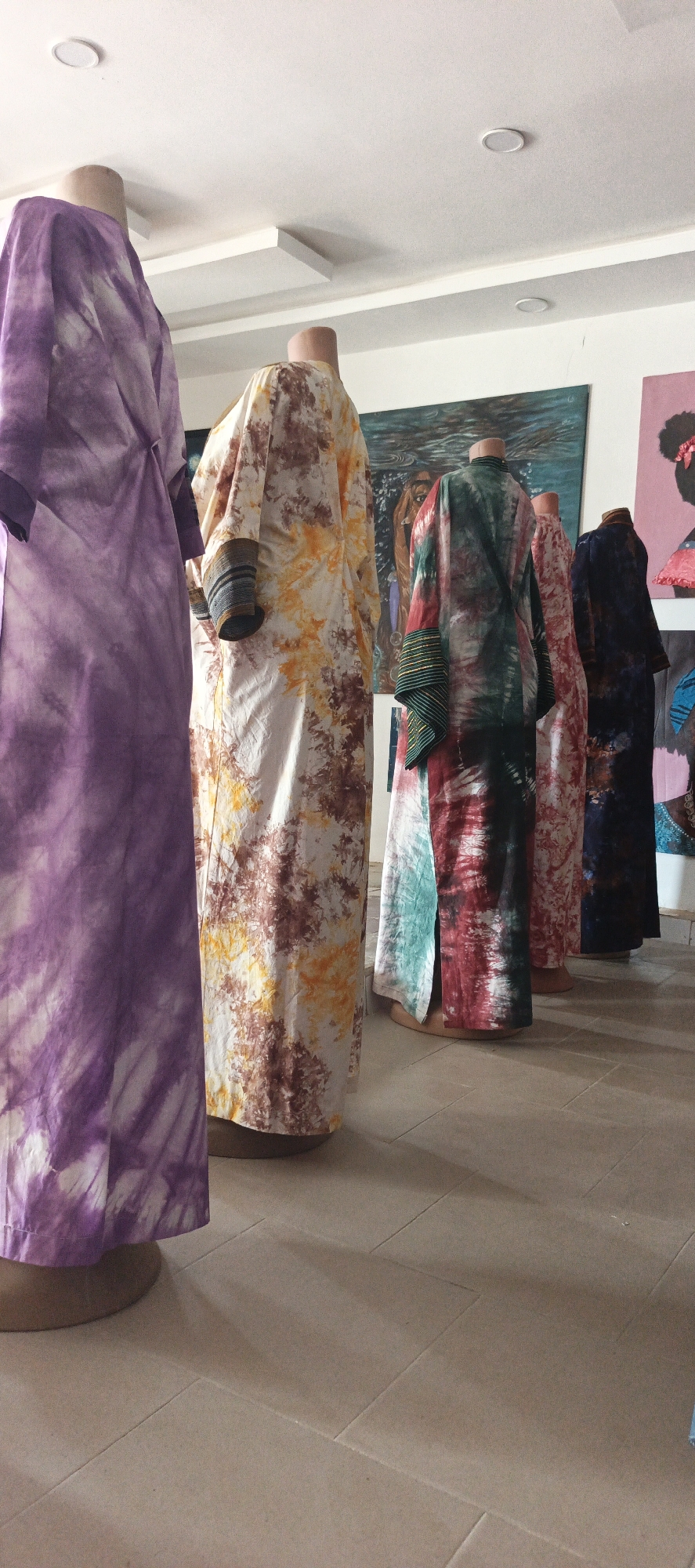 The image displays an indoor scene, likely a shop or gallery, featuring five headless mannequins adorned with long, flowing tie-dye robes or dresses. The mannequins are arranged in a diagonal line, extending from the foreground on the left to the background on the right, creating a sense of depth.
The robes exhibit a variety of vibrant tie-dye patterns and colors. The mannequin closest to the viewer on the left wears a prominent purple and white garment. The next mannequin showcases a robe with a blend of yellow, orange, brown, and white, with distinctive striped cuffs. The central mannequin is dressed in a striking red, green, and black tie-dye robe, also featuring striped cuffs. Behind it, another mannequin wears a red and white patterned dress, and the furthest one on the right is clad in a dark blue or purple and white garment. These garments appear to be traditional or culturally inspired attire, possibly African boubous.
The background reveals several pieces of artwork displayed on white walls. To the left, a large painting with blue and green hues, possibly depicting abstract figures or water, is visible. To its right, a pink painting features a silhouette of a person's head, suggesting an artistic representation of a woman, possibly with an afro hairstyle. Other less distinct artworks can be seen further in the background.
The ceiling is white and features recessed circular lights and rectangular panels, suggesting a modern interior. The floor is covered with light-colored tiles, casting subtle shadows from the mannequins and their bases. The overall lighting is bright, indicating it is likely daytime. There is no visible text in the image. The setting suggests an exhibition or sale of handcrafted garments and art, consistent with a cultural or artistic venue in Akwanga, Nigeria.