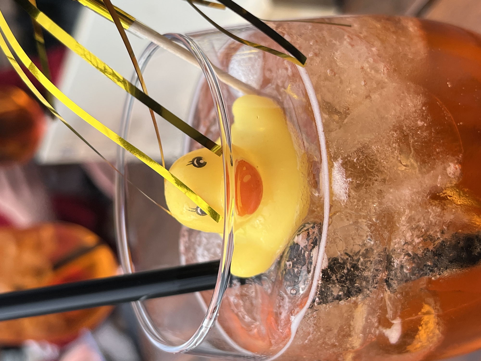 A close-up, high-angle shot shows a glass filled with an orange liquid and ice, featuring a yellow rubber duck partially submerged within. The glass is adorned with gold and black tinsel-like decorations. A black straw is visible, partially obscuring a blurred, orange object in the background, potentially a pumpkin. The image may have been taken in Cochem, Germany, though no specific architectural or environmental details confirm its location.