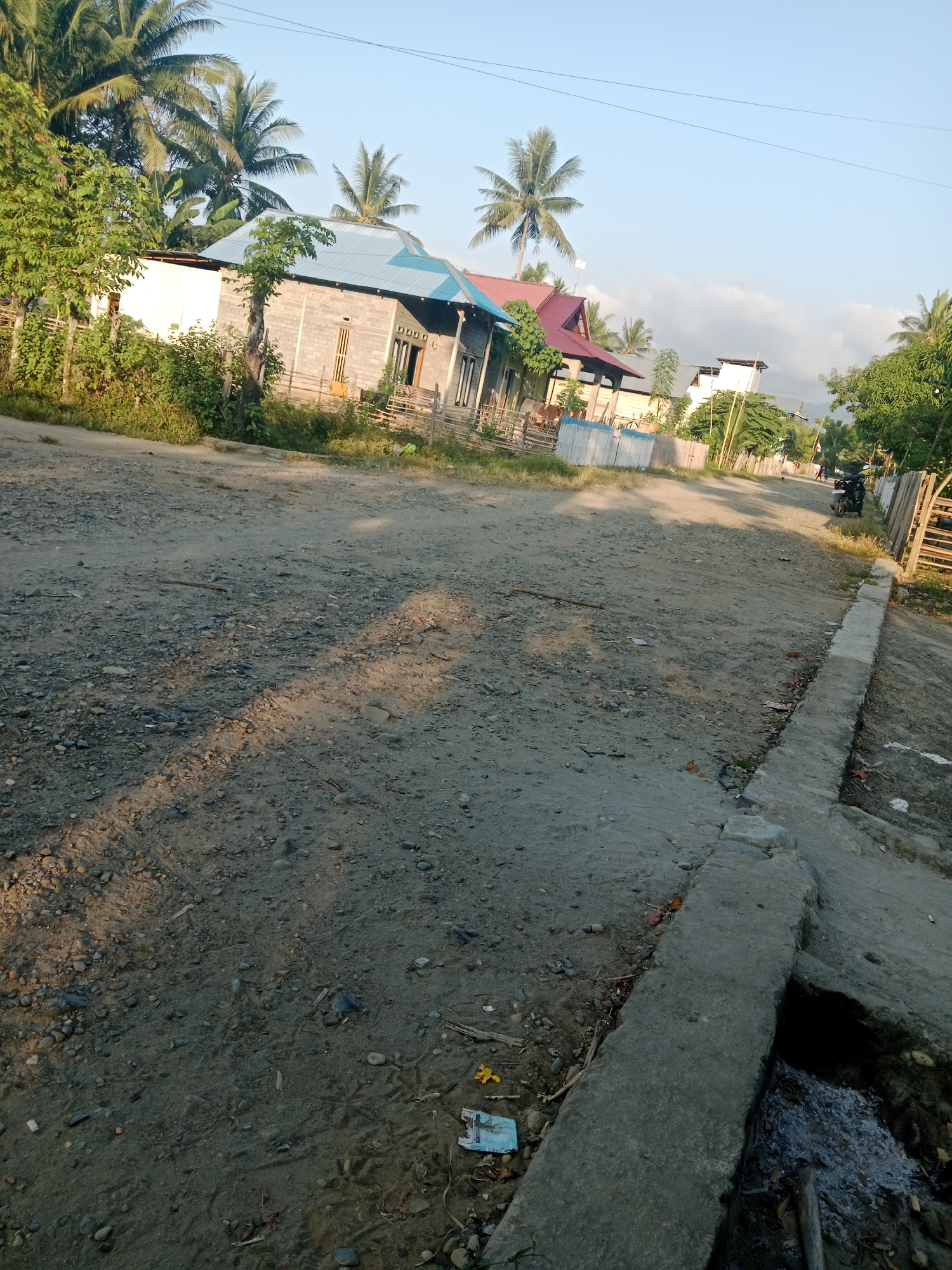 A dirt road curves through a village in Lakea Satu, Indonesia. On the left, houses with pale walls and corrugated metal roofs are partially visible behind a wooden fence and lush green foliage. Coconut trees with their fronds reach skyward. On the right, the road is bordered by a low concrete barrier that follows its course. Beyond the barrier, a few more houses and trees are scattered along the roadside. A dark-colored motorcycle is parked on the right side of the road near a wooden fence. The ground is covered in dirt, small stones, and scattered debris. The sky is a clear, light blue with a few faint clouds. The sun casts long shadows across the road, indicating it is either early morning or late afternoon.