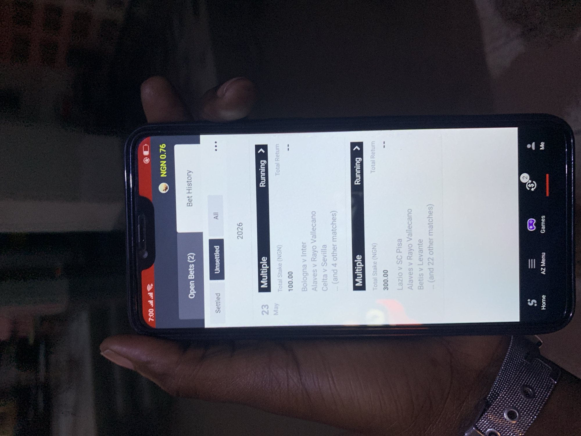 A person is holding a mobile phone displaying a betting app. The phone screen shows two open bets, one with a total stake of 100 NGN on a multiple bet including Bologna v Inter, Alaves v Rayo Vallecano, and Celta v Sevilla, and another with a total stake of 300 NGN on a multiple bet including Lazio v SC Pisa, Alaves v Rayo Vallecano, and Betis v Levante. The app indicates "Running" status for both bets. The phone displays a 7:00 AM time and a balance of 0.76 NGN. The person's hand and part of their watch are visible. The environment is dark and out of focus.
